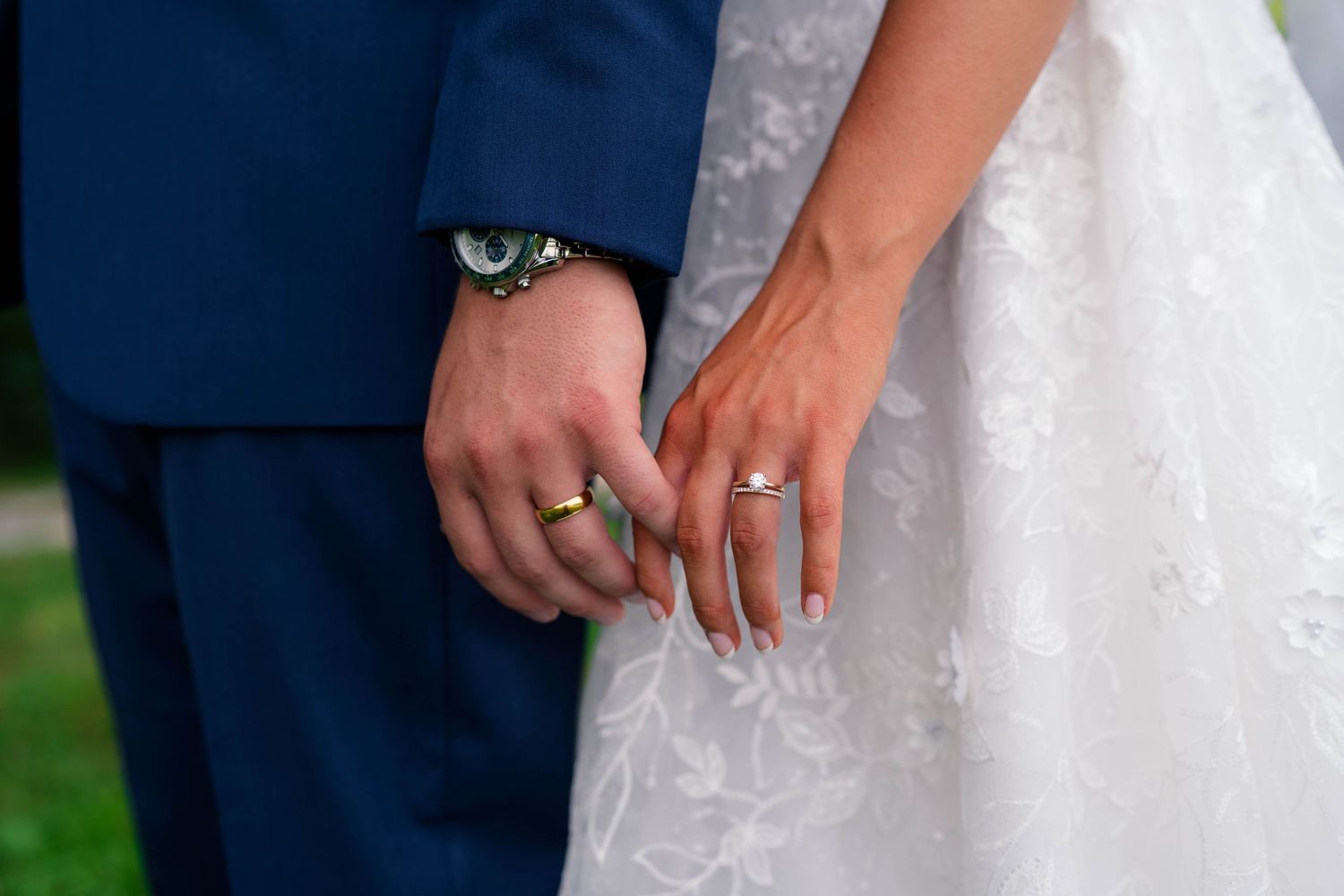 Close-up of bride and groom holding hands showing wedding rings, lace gown and navy suit.