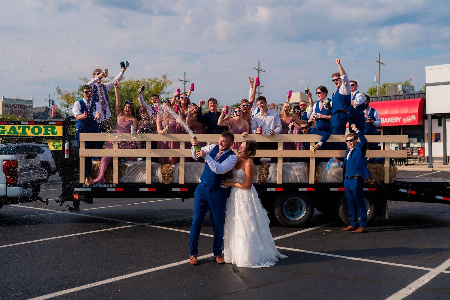 Bride and groom kiss in parking lot while wedding party celebrates on decorated flatbed truck behind them.