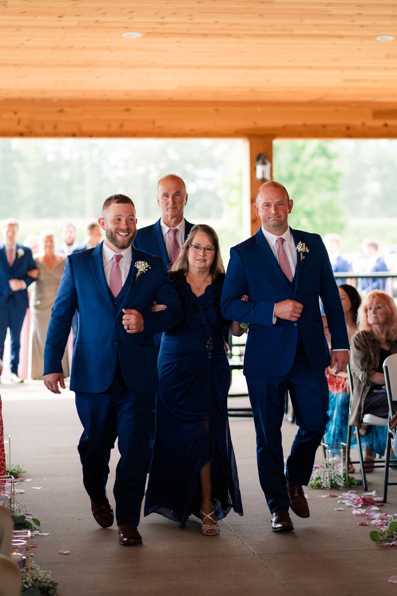 Woman escorted down wedding aisle by two groomsmen in navy suits under wooden pavilion.
