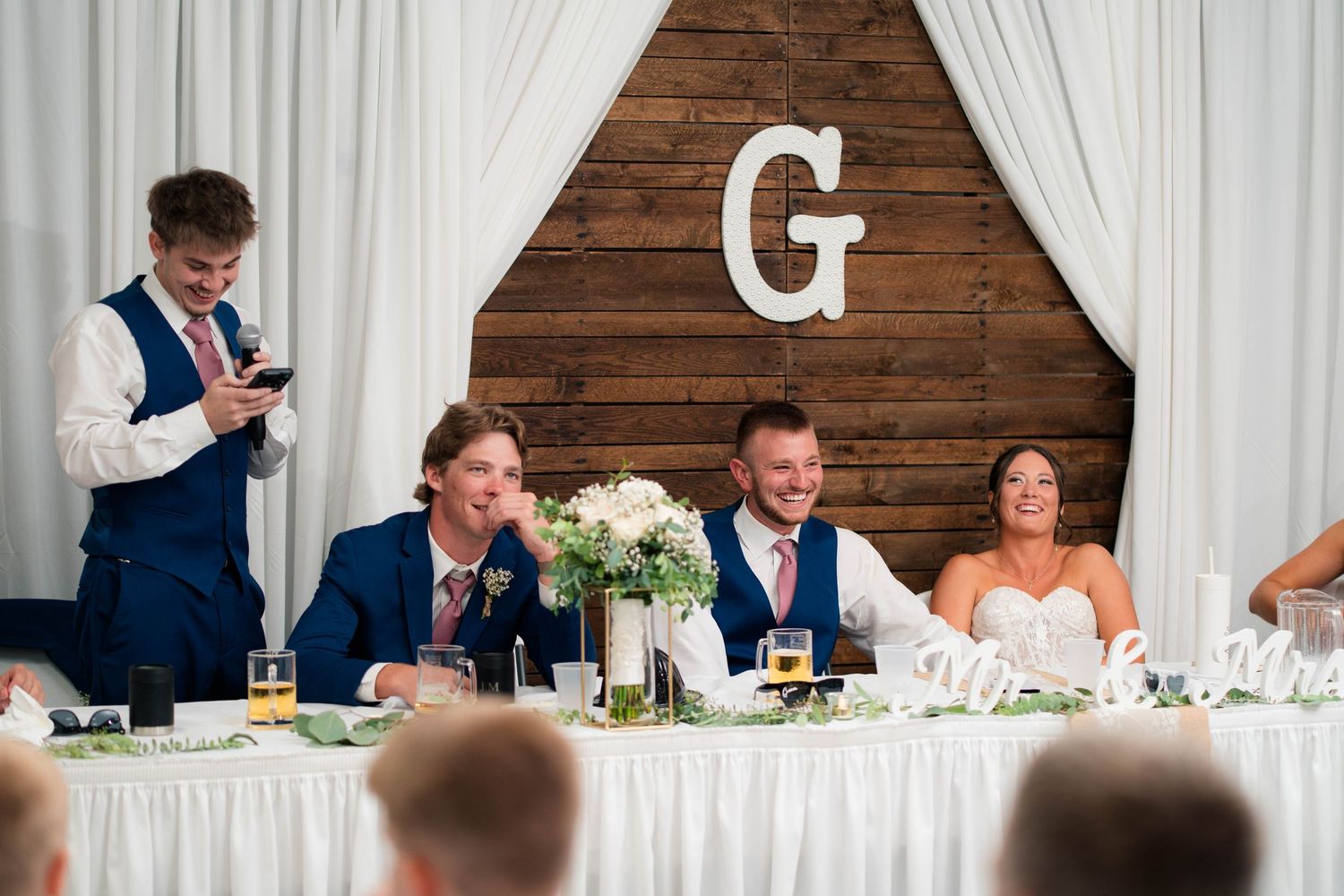 Wedding party laughs at sweetheart table during reception speech with rustic wooden G monogram backdrop.