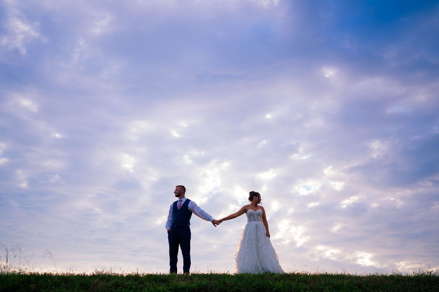 Bride and groom hold hands on grassy hill under dramatic purple cloudy sky at sunset wedding photo.