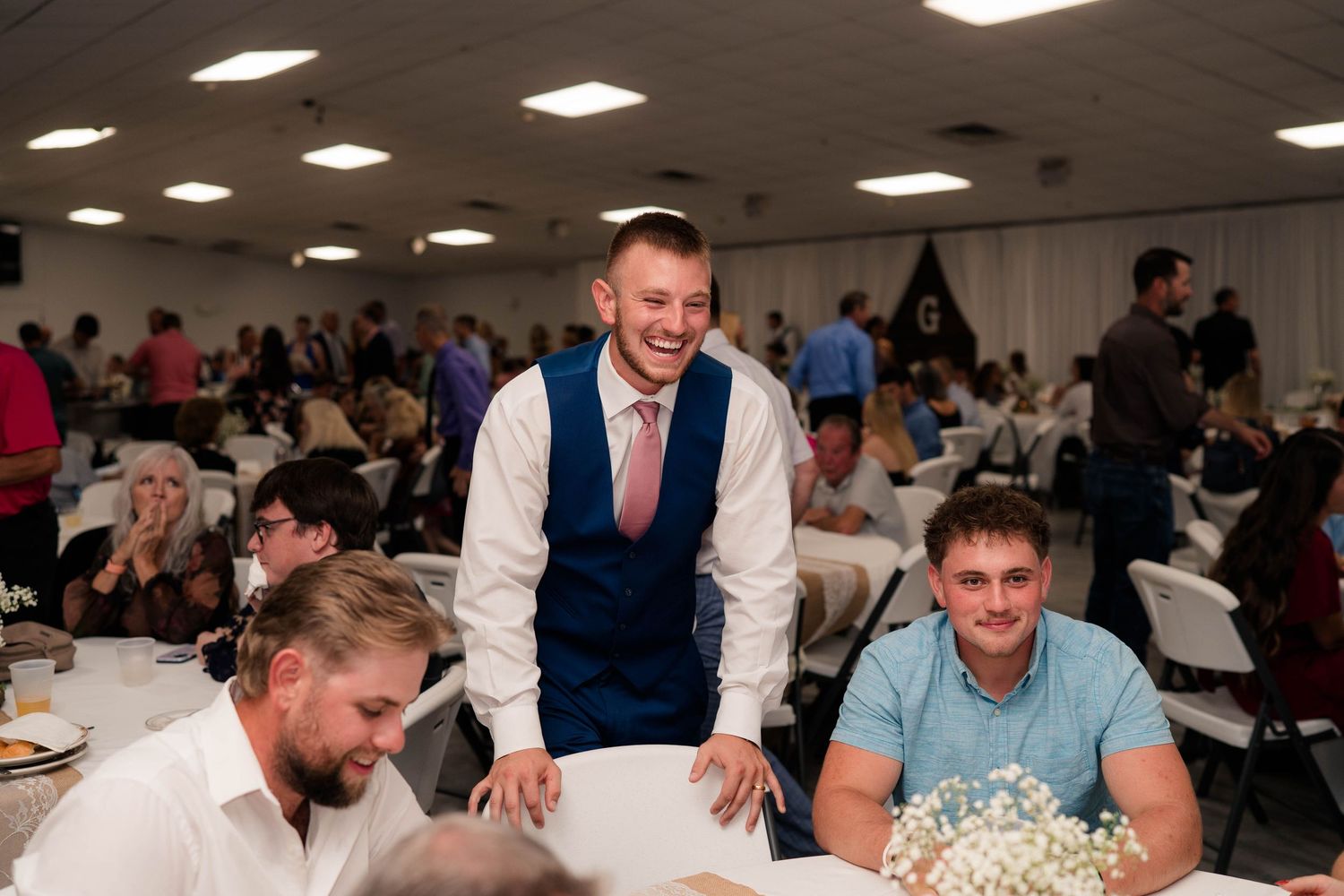 A smiling groom in a navy vest and pink tie laughs with guests at a wedding reception dinner.
