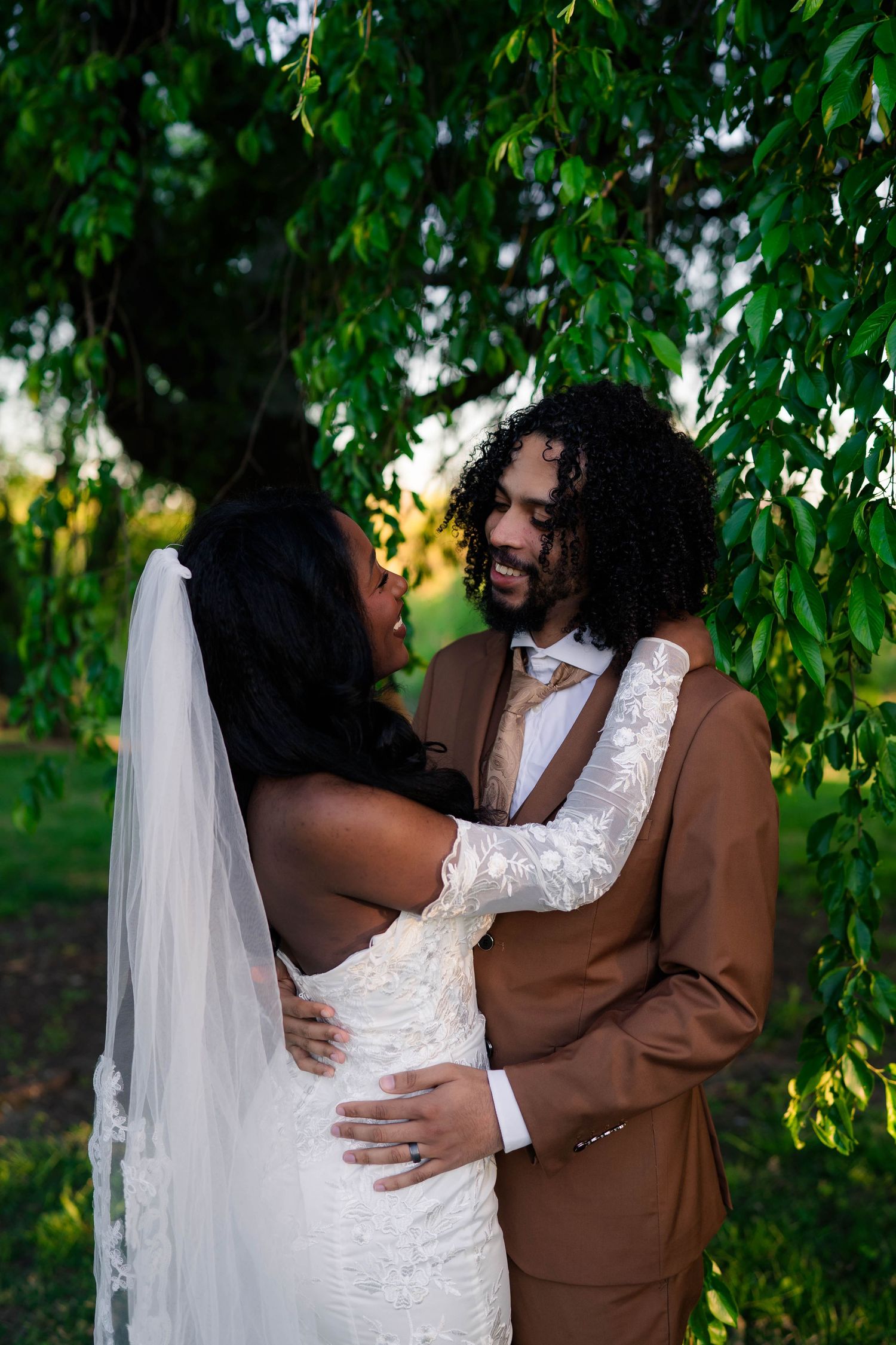 A bride and groom embrace under a weeping willow tree, wearing white lace gown with veil and brown suit.