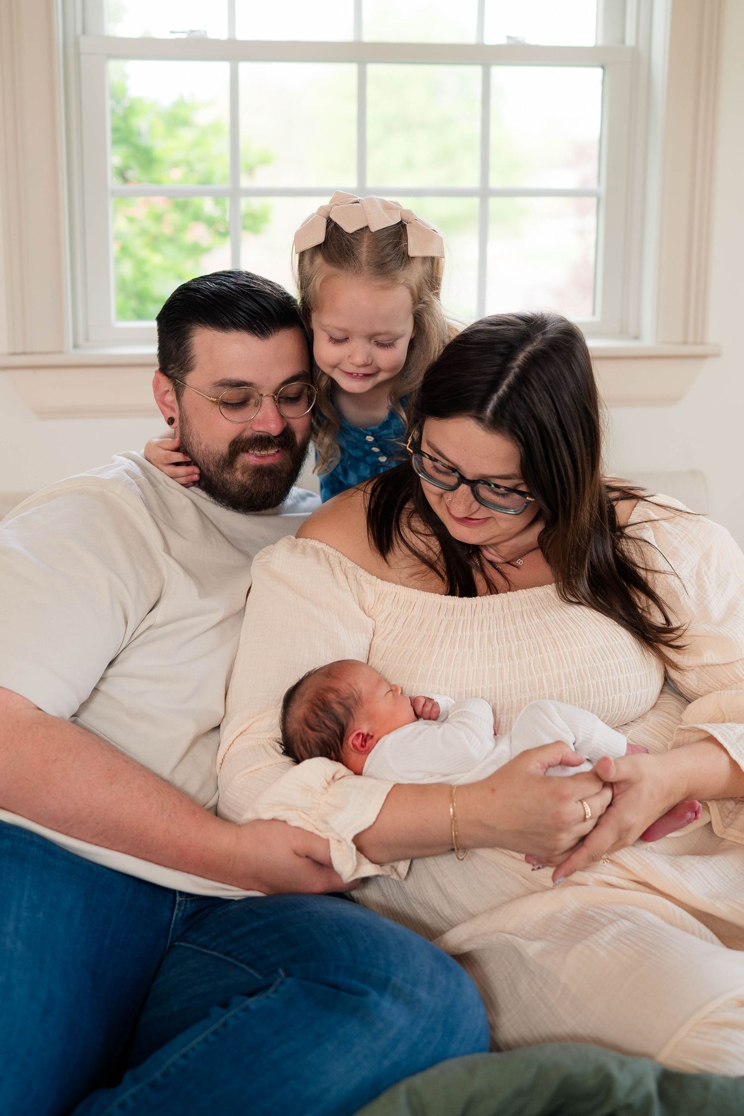 Family of four admiring their newborn baby, parents and toddler girl gathered together near a bright window.