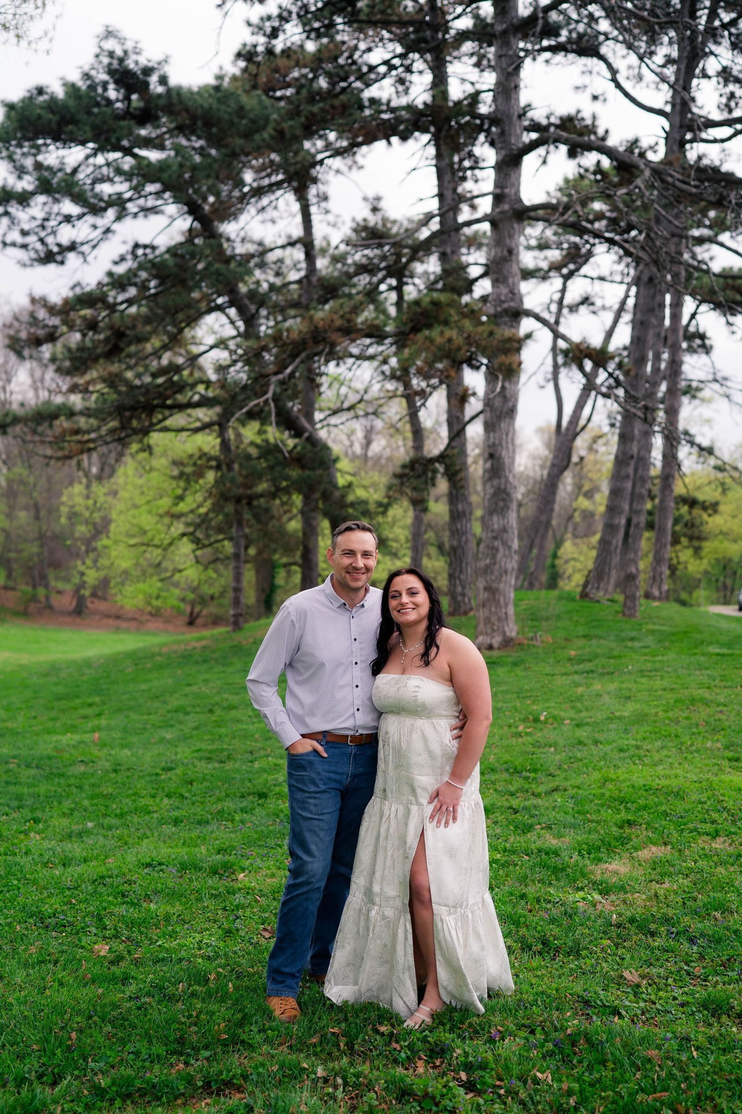 A smiling couple poses together on a grassy park lawn with tall pine trees in the background.