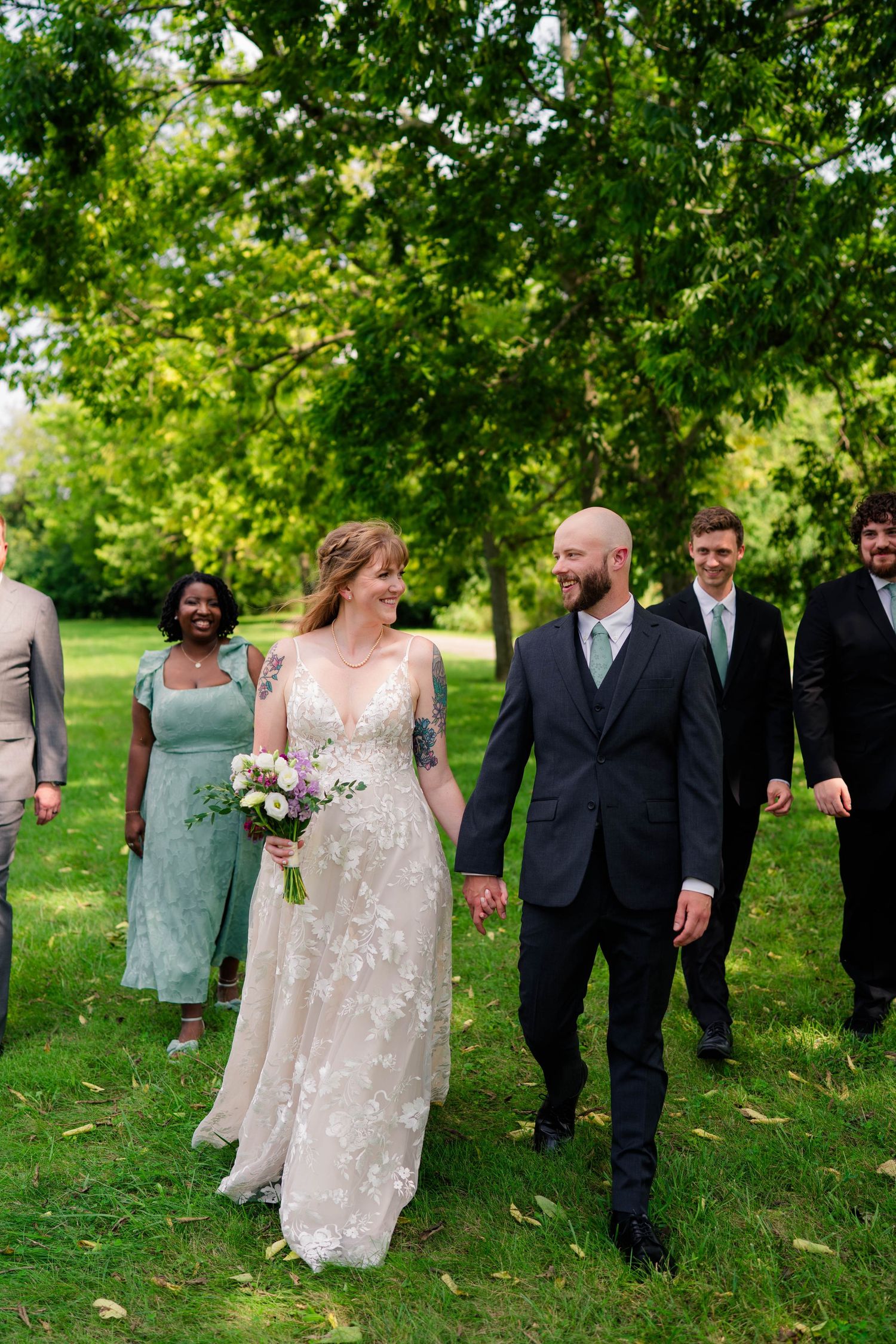 Bride and groom hold hands walking with wedding party through lush green park setting.