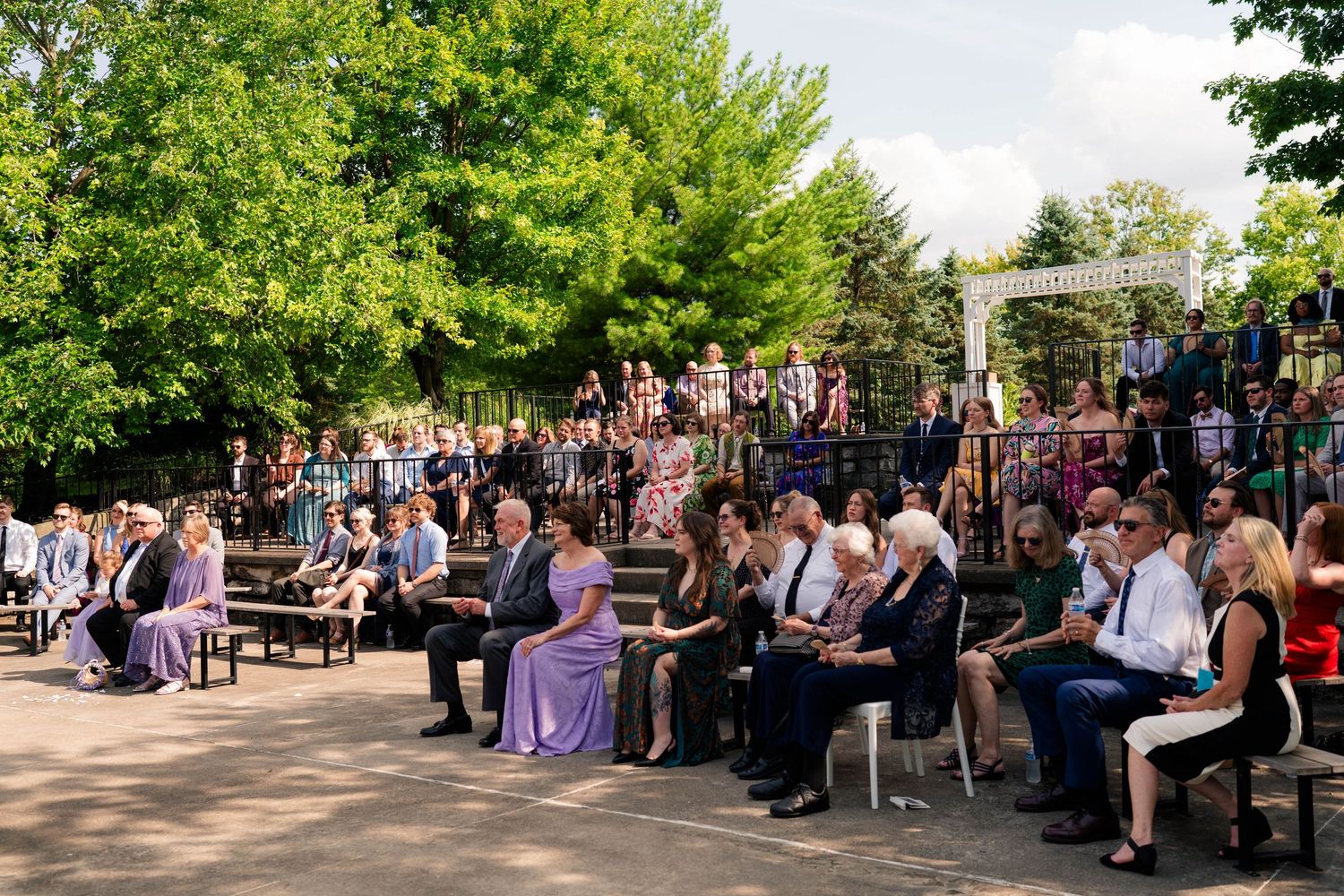 Guests seated outdoors in sunlit garden setting during an elegant wedding ceremony.