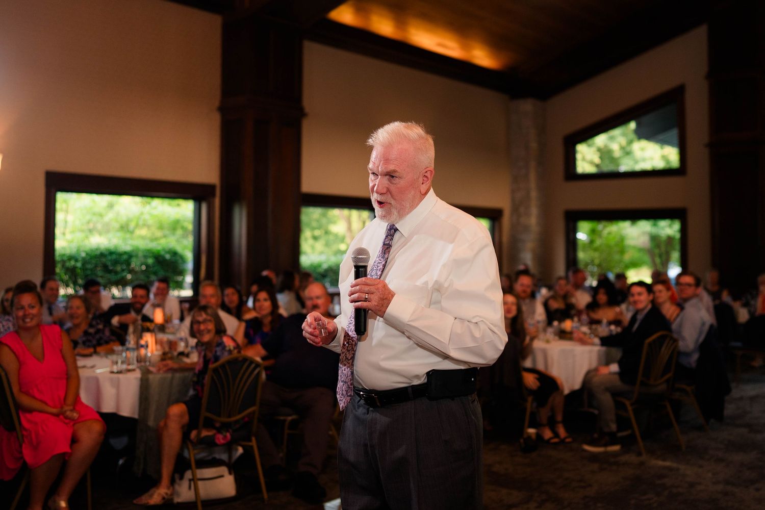 Elderly man giving a speech with microphone at a crowded wedding reception indoors.