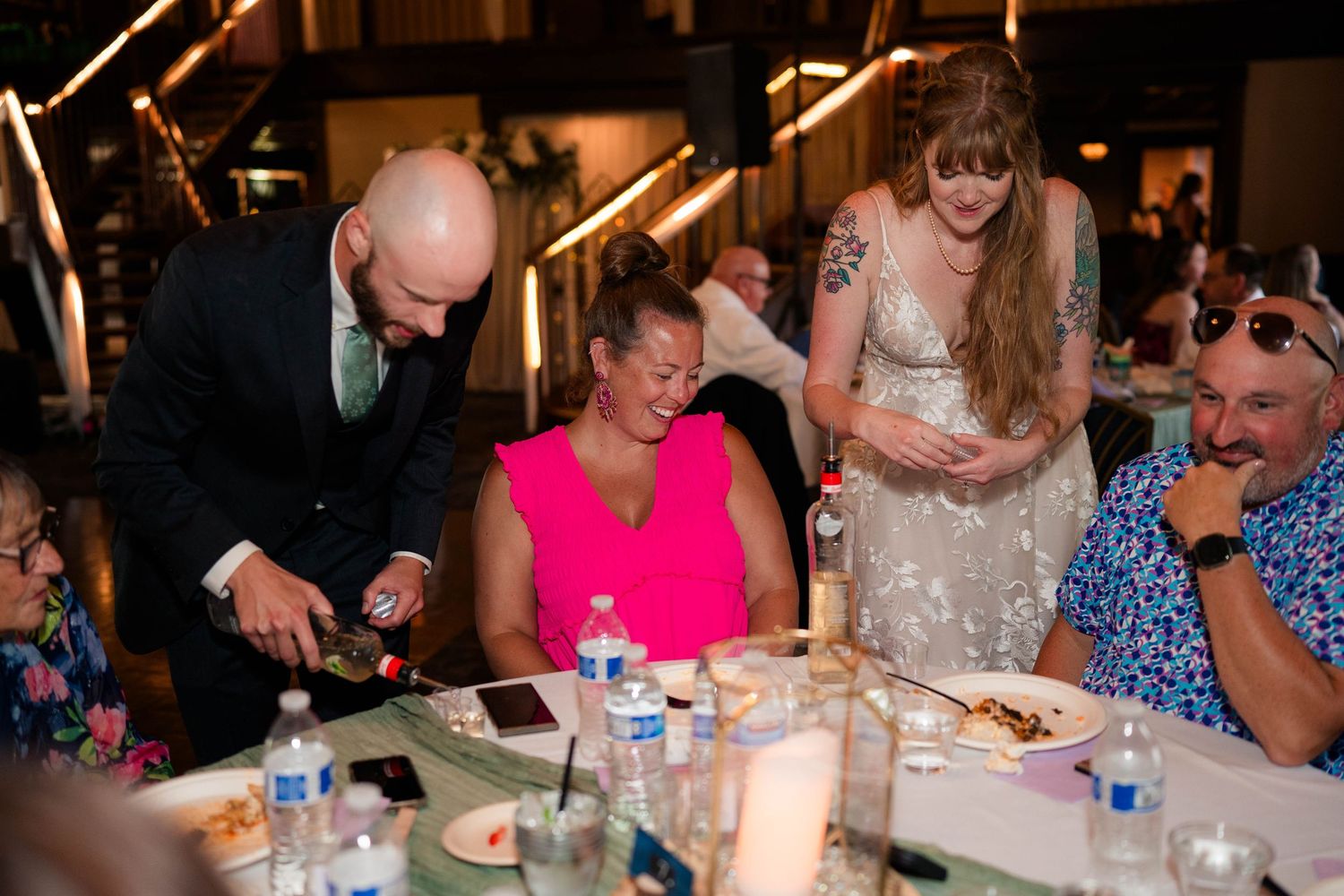 Bald groom pours wine for guest in pink dress at wedding reception dinner table.