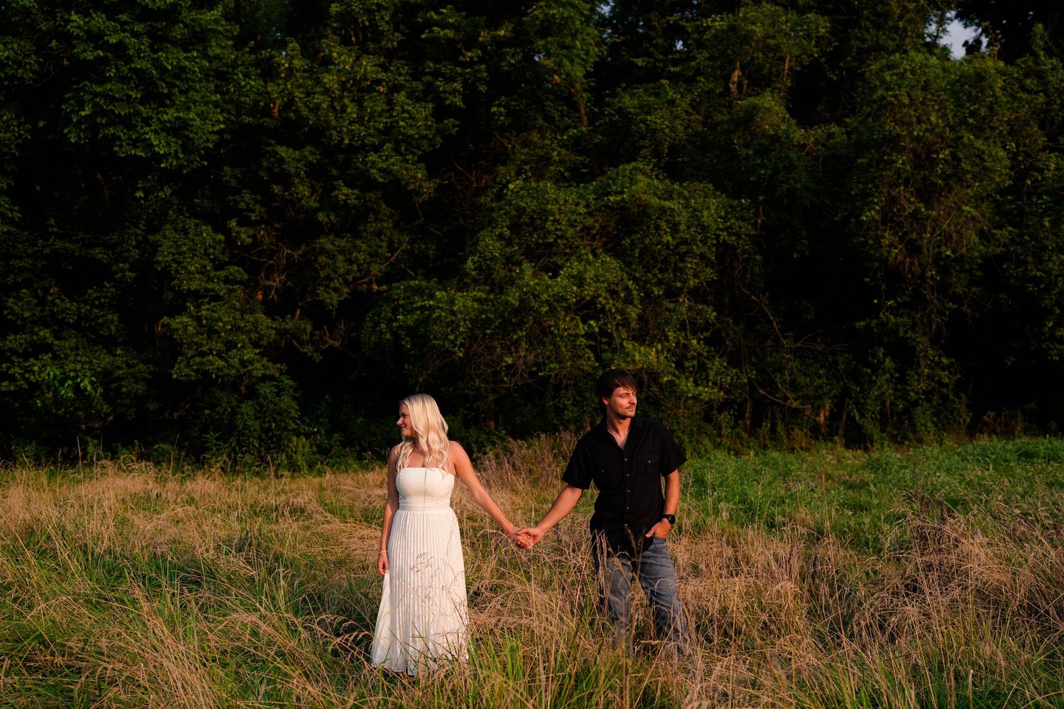 Couple holding hands in golden field with lush green forest backdrop during sunset engagement photo session.
