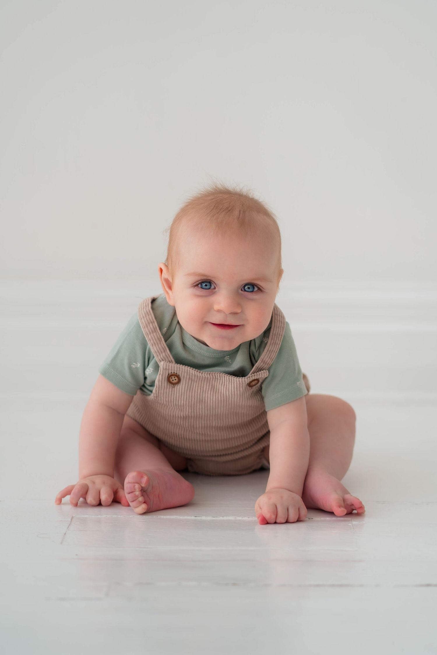 Smiling baby in green onesie and tan overalls sitting on white floor.