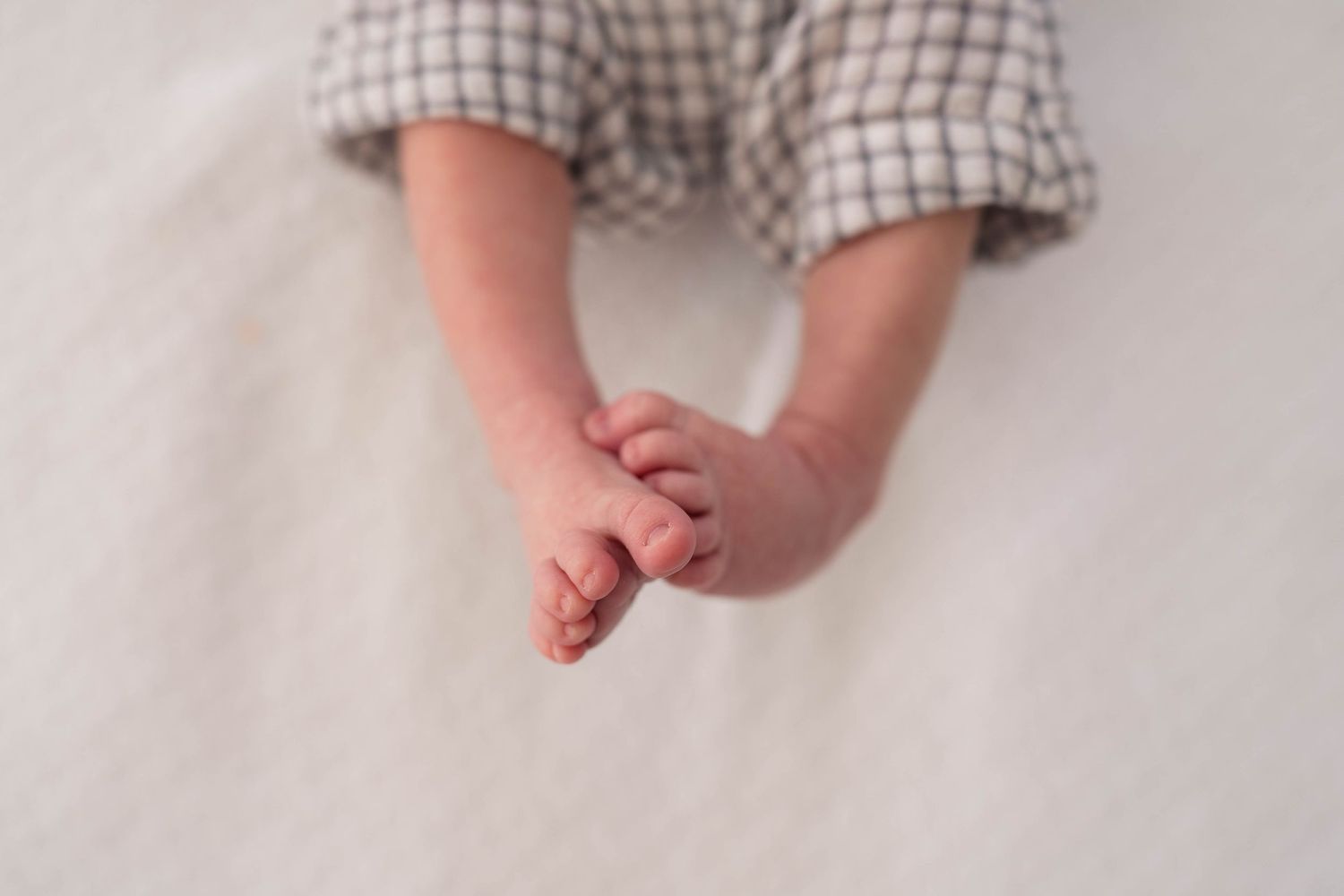 Close-up of a newborn baby's tiny hands and feet in a plaid onesie on a white blanket.