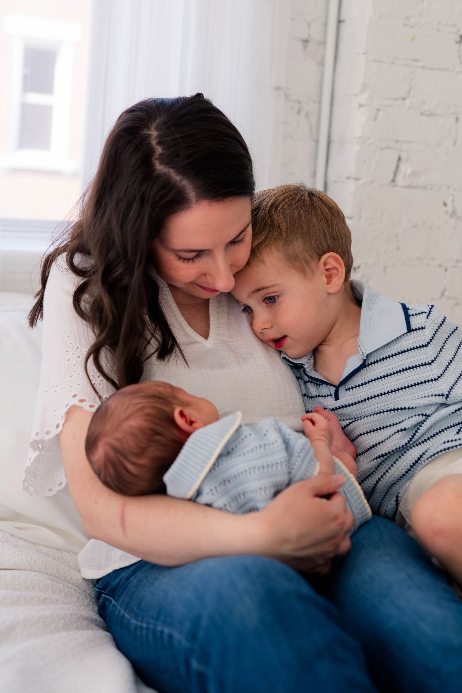 Brunette mother sitting on bed with toddler son and newborn baby, family bonding moment with siblings.