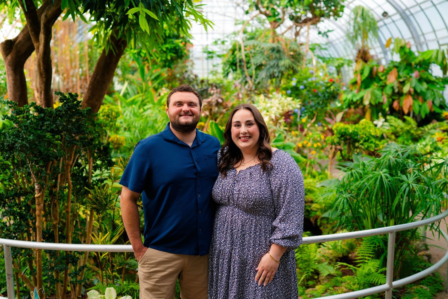 Couple smiling together inside a lush tropical greenhouse conservatory with vibrant green plants.