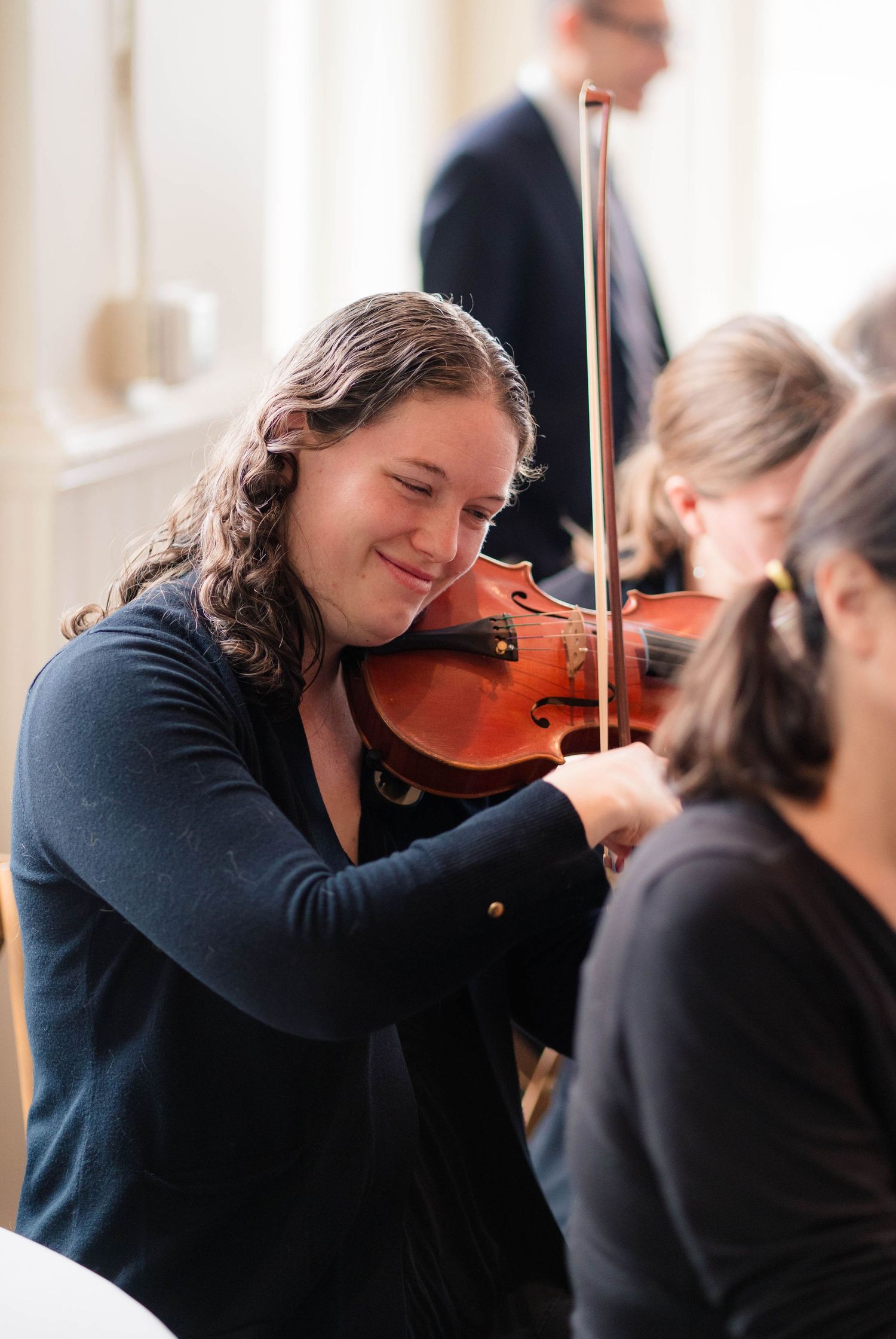 Smiling young woman plays violin at an elegant indoor event with natural window lighting.