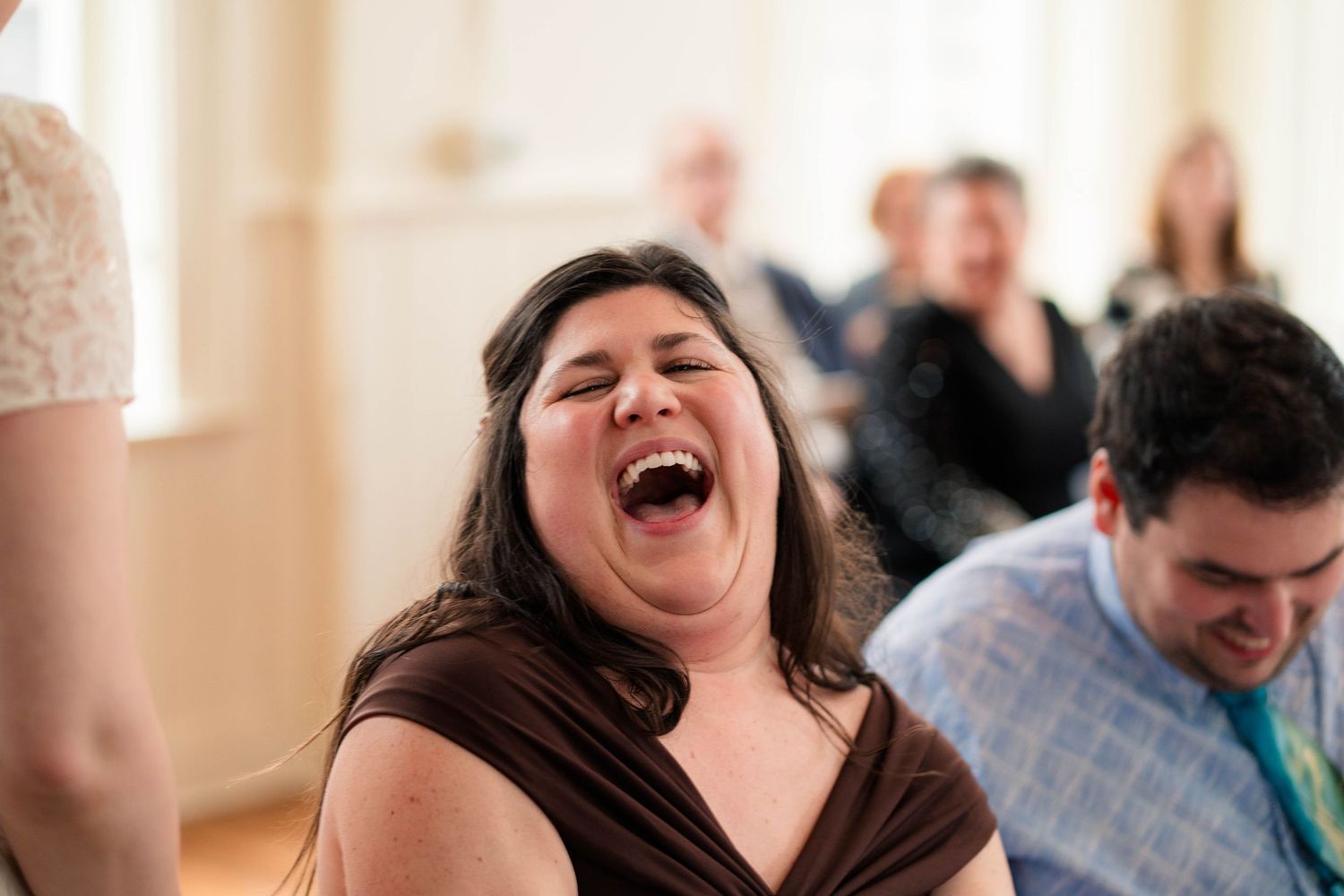 Woman laughing loudly at wedding reception with guests smiling and celebrating in the background.