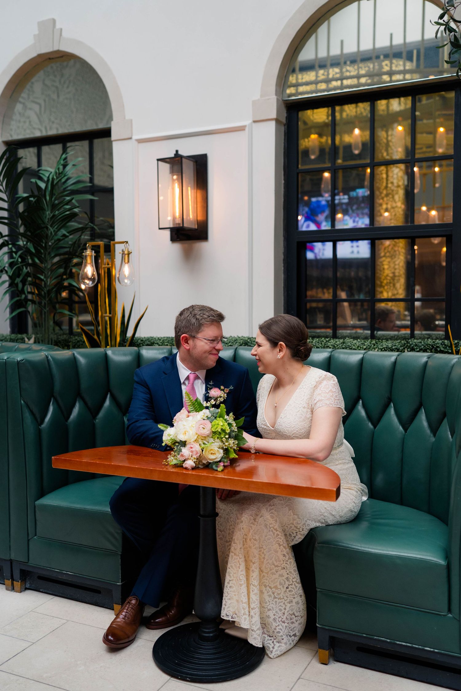 Bride and groom sit together in green leather booth, sharing an intimate moment with bouquet.