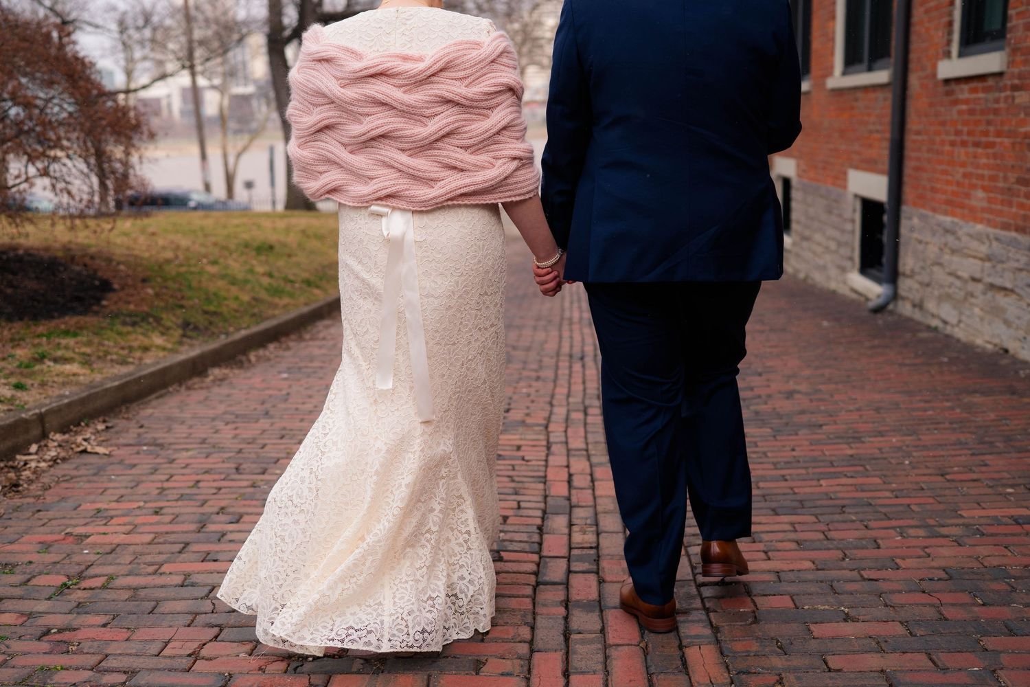 Rear view of bride in lace gown with pink cable-knit wrap and groom in navy suit walking on a brick path.