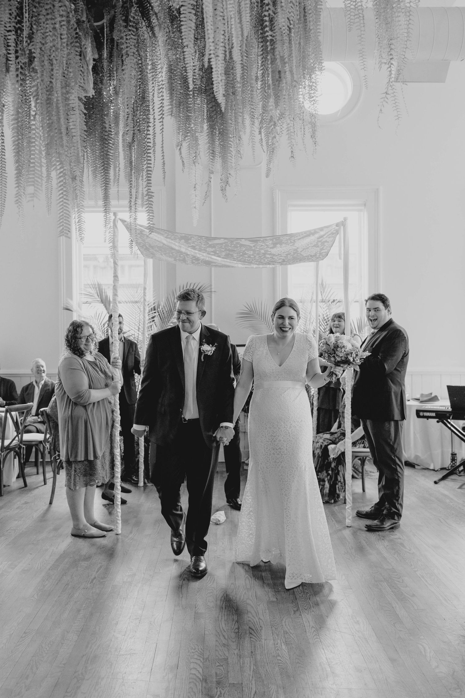 Black and white photo of joyful newlyweds walking down aisle after indoor wedding ceremony.