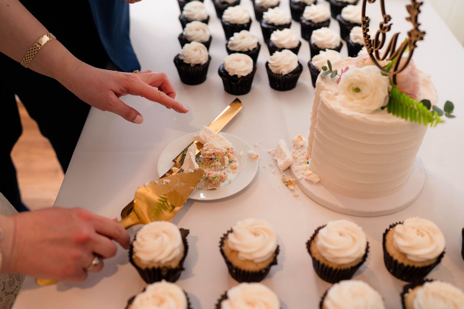 Close-up of hands cutting white wedding cake with gold knife beside rows of frosted cupcakes.