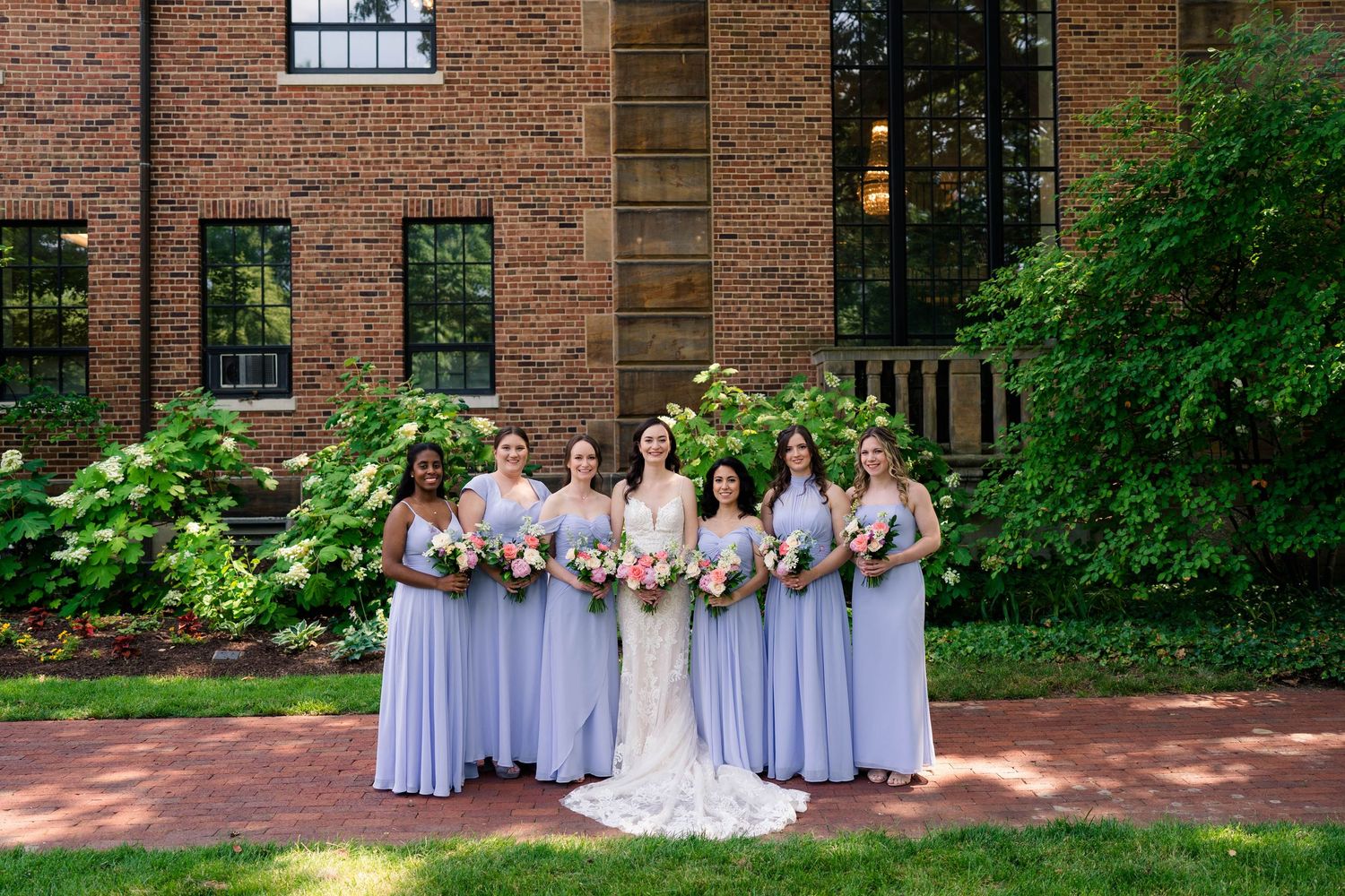 Bride with bridesmaids in lavender gowns holding bouquets outside a brick building.