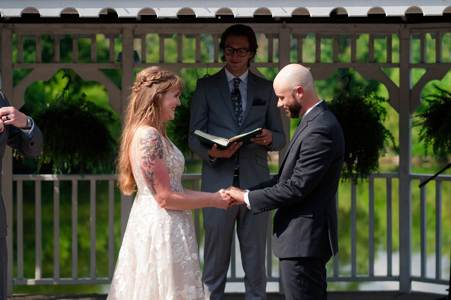 Bride and groom hold hands during outdoor wedding ceremony while officiant reads from book at decorated gazebo.