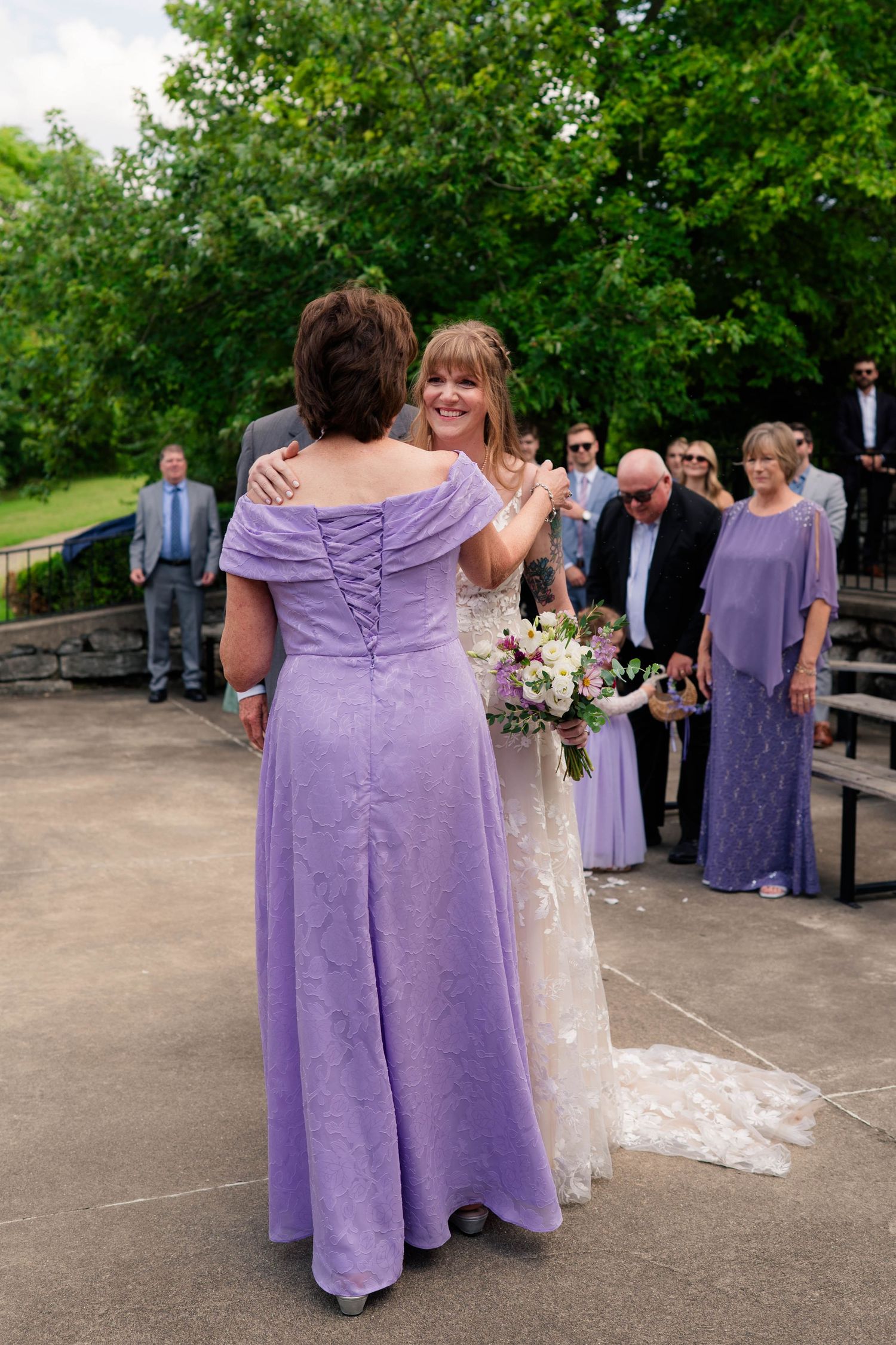 Bride embraces woman in lavender gown during outdoor wedding ceremony.
