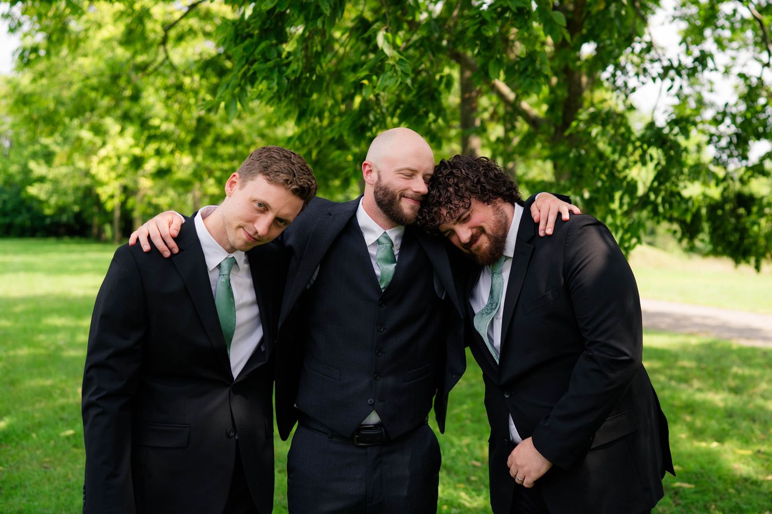 Groom and two groomsmen in black suits laughing together in a park setting.