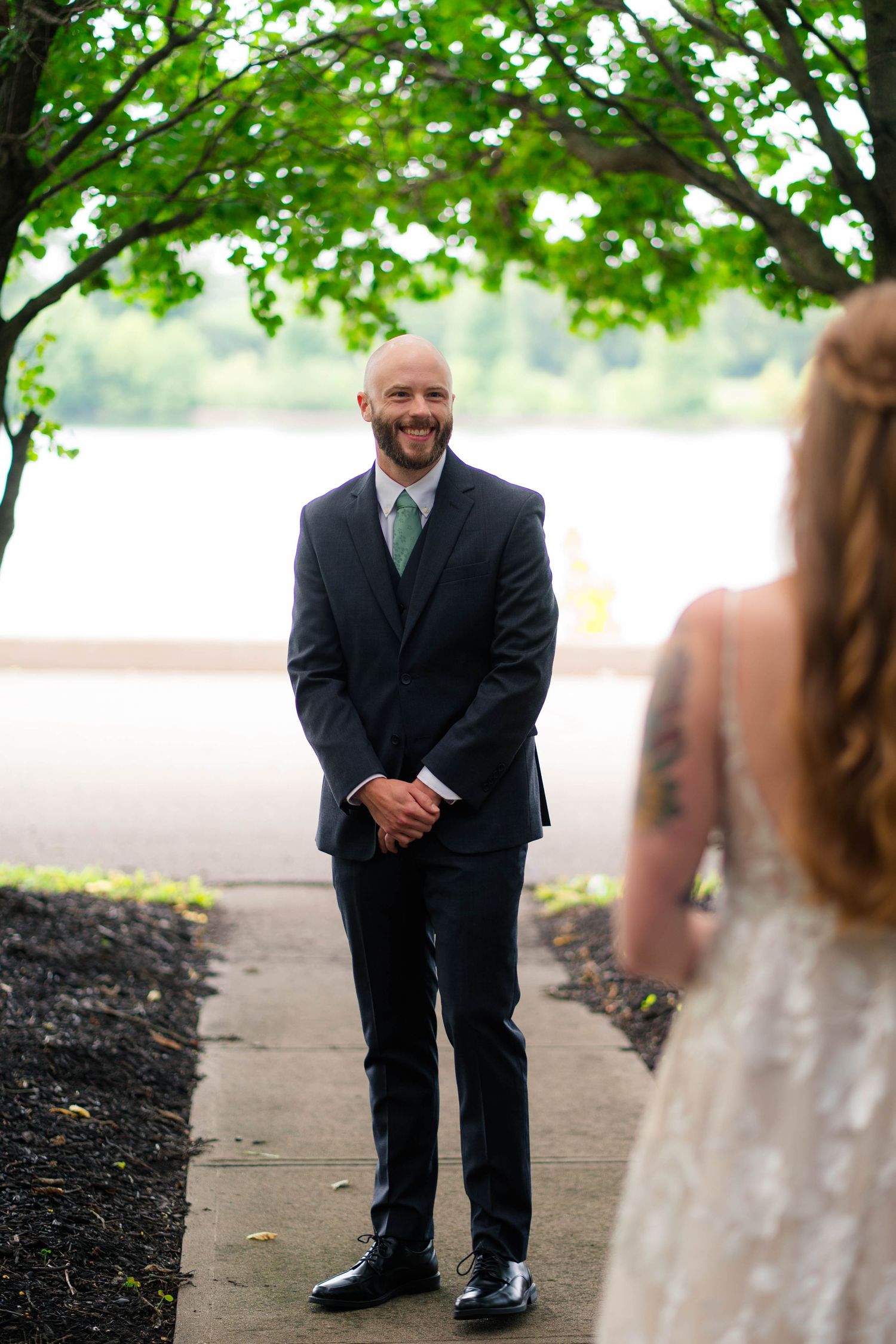 Emotional groom in navy suit smiling at first look with tattooed bride in lace wedding dress outdoors.