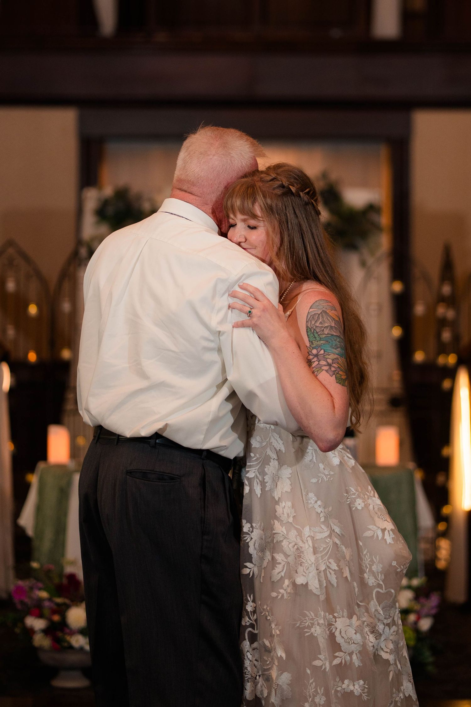 Bride with floral tattoo shares emotional father-daughter dance at candlelit wedding reception.