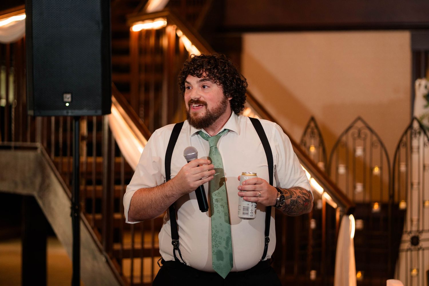 Curly-haired groomsman with suspenders and green tie gives heartfelt toast at wedding.