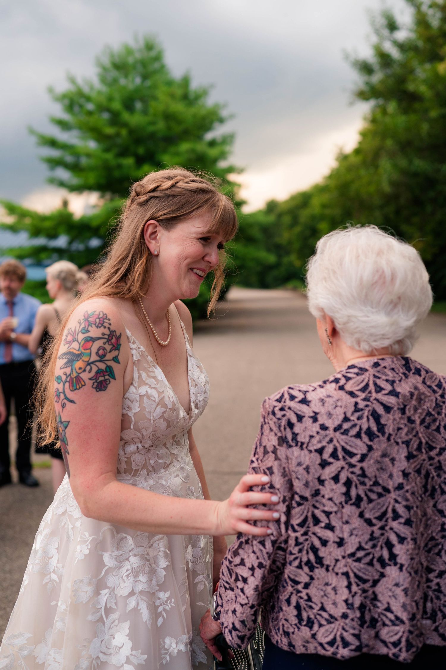 Bride in lace gown with floral arm tattoo smiles warmly at elderly guest outdoors.