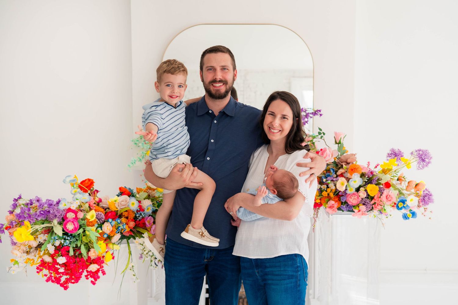 Happy family with toddler and newborn posing with colorful floral arrangements.