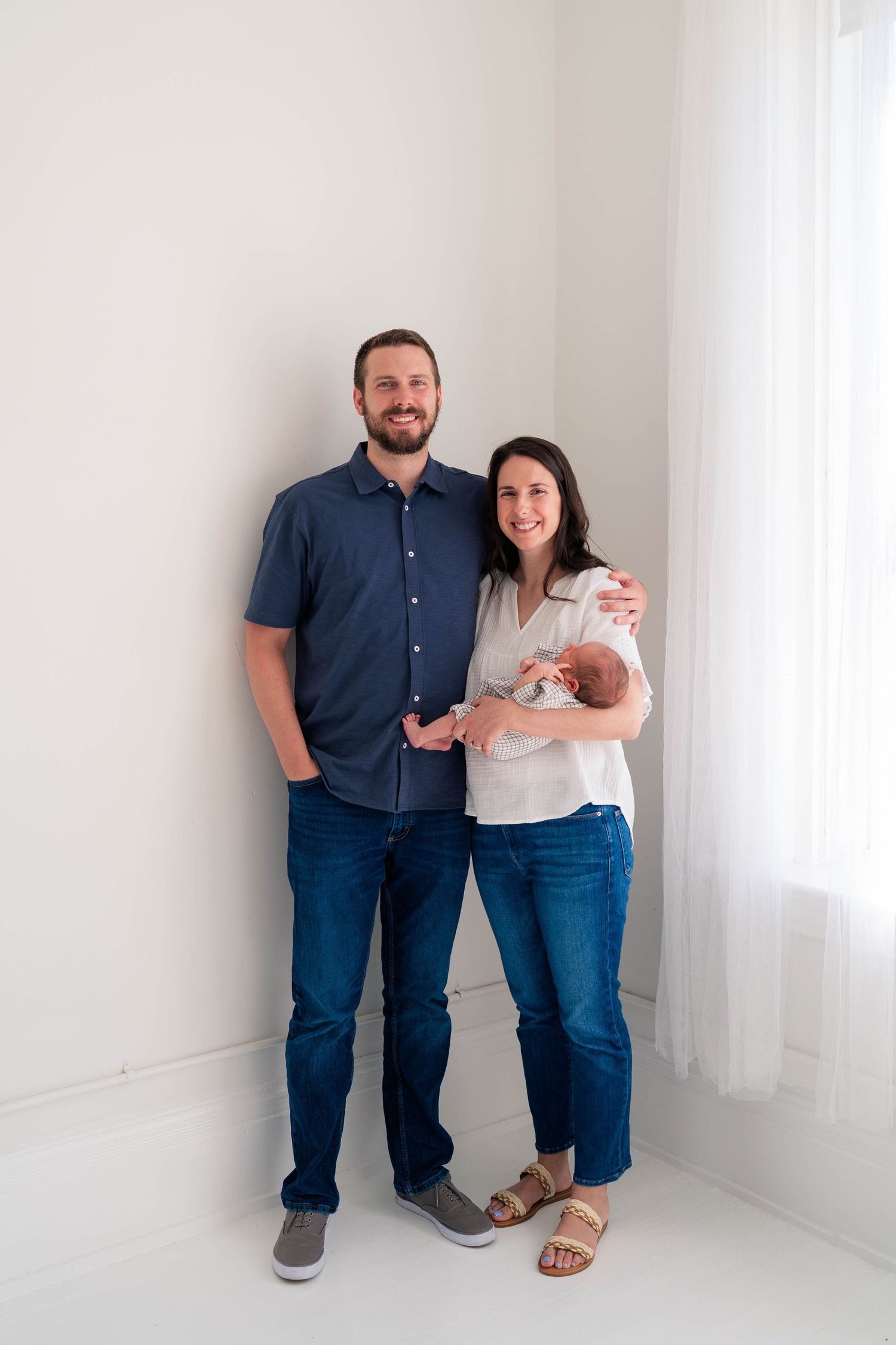 Happy couple in casual attire smiling while holding their newborn baby in a bright white studio room.