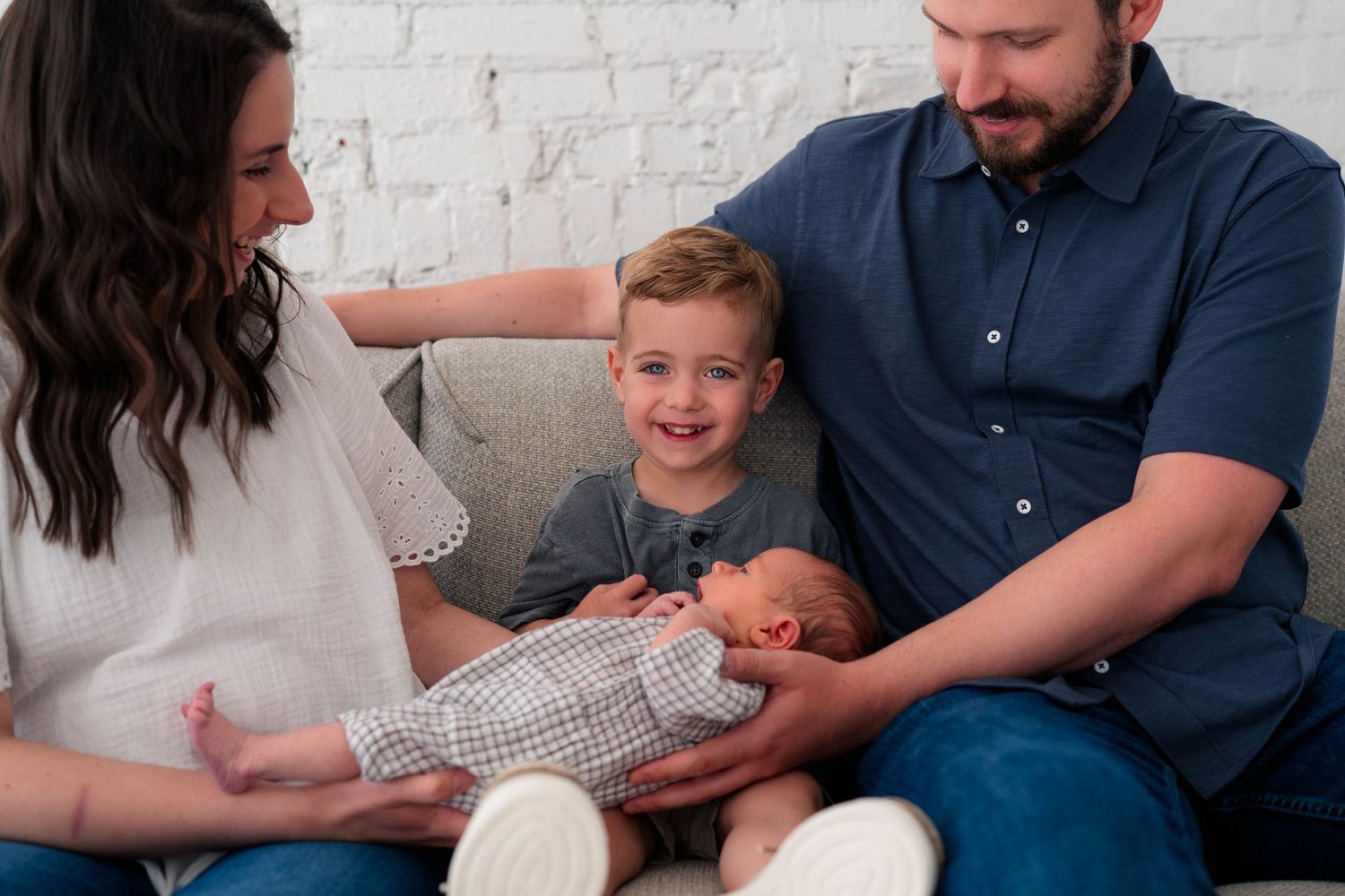 Happy family with parents, toddler boy, and newborn baby sitting together on a couch for a professional photo session.