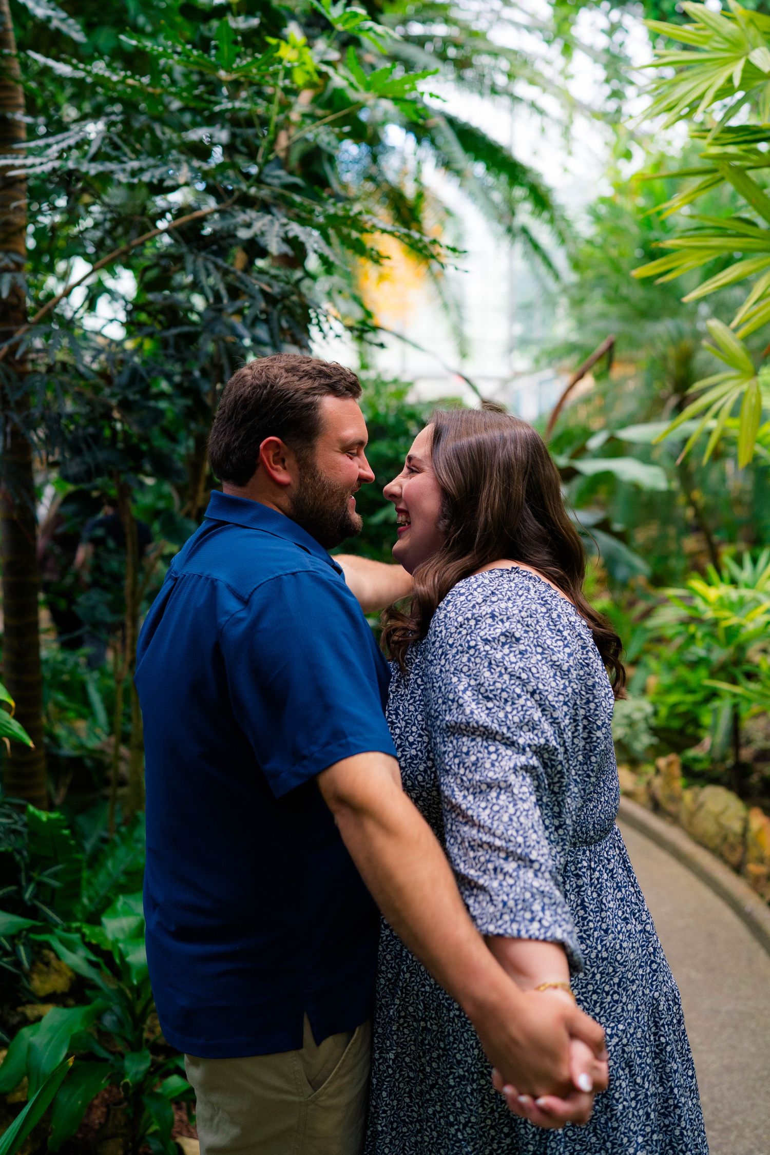 A joyful couple holds each other close, laughing together amid vibrant tropical plants in a greenhouse.