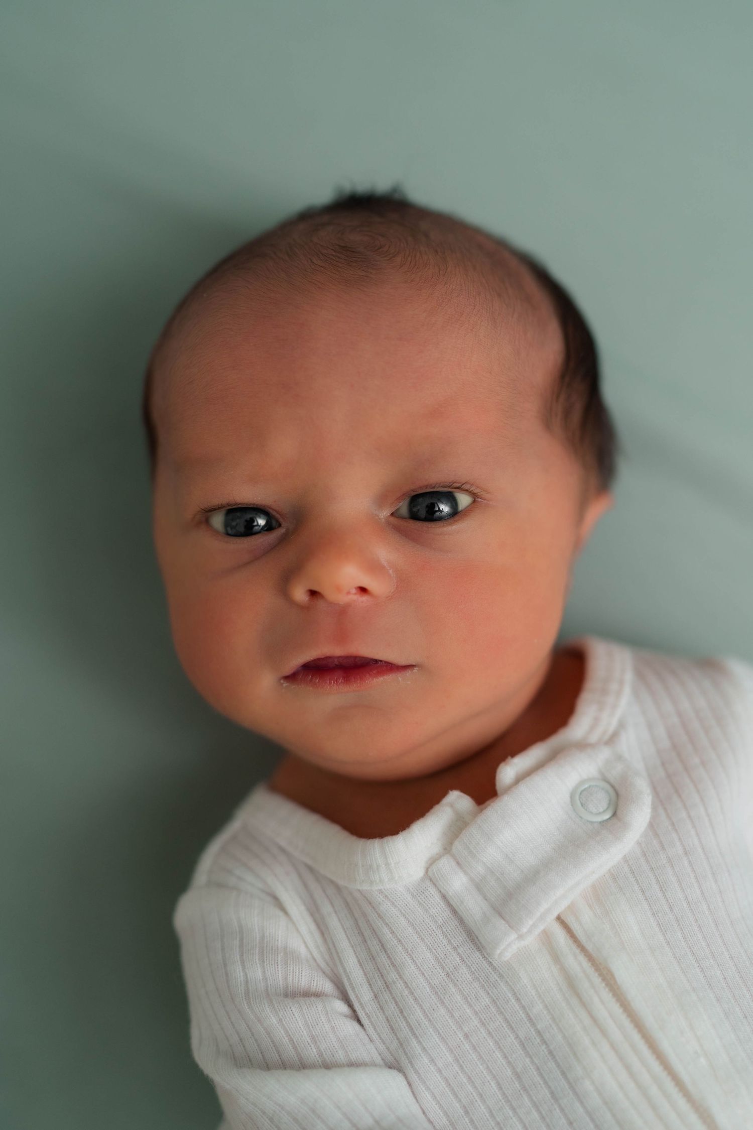 Alert newborn baby with dark eyes in white ribbed onesie on green background.
