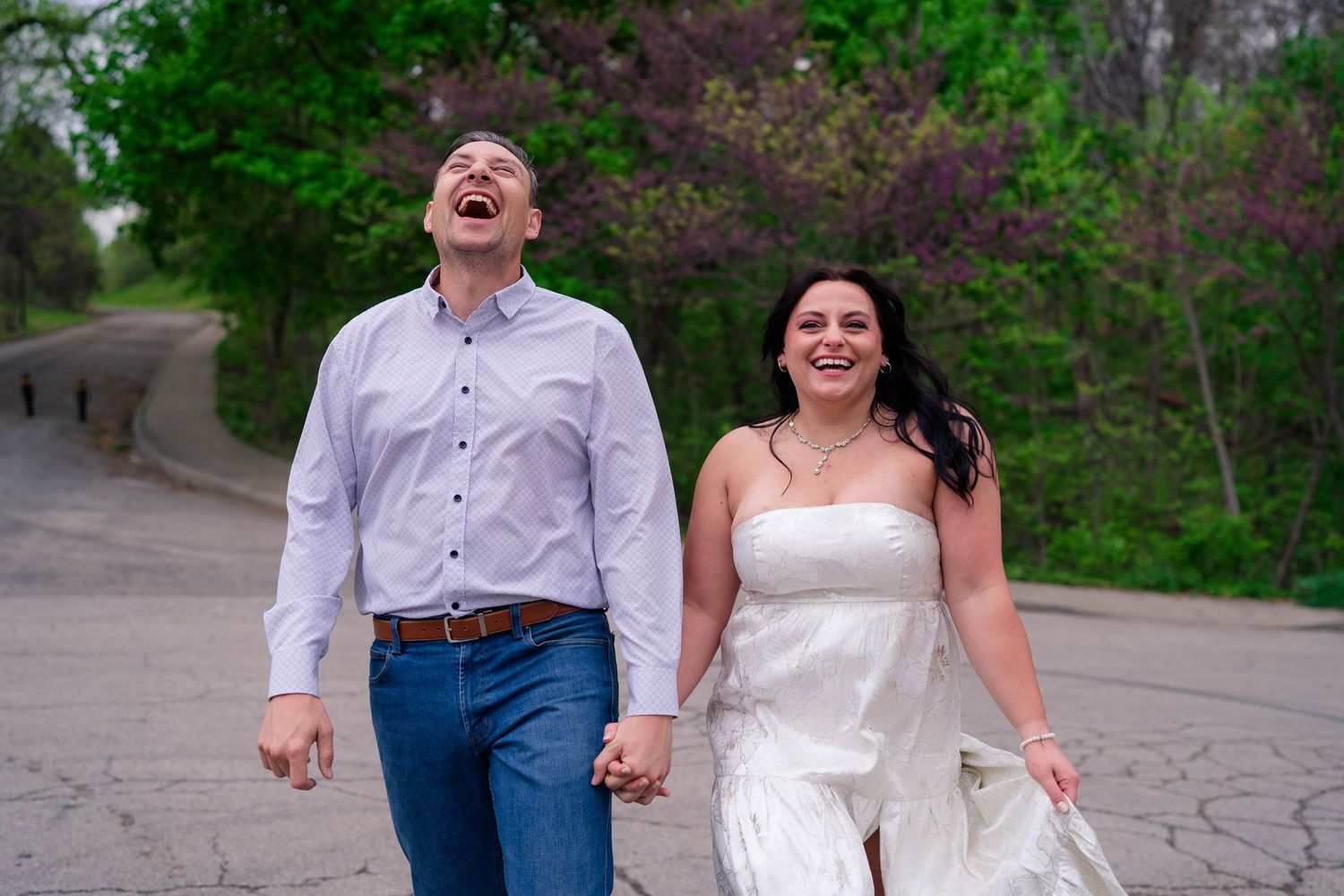 Happy couple laughing and holding hands while walking down a tree-lined path during engagement photos.