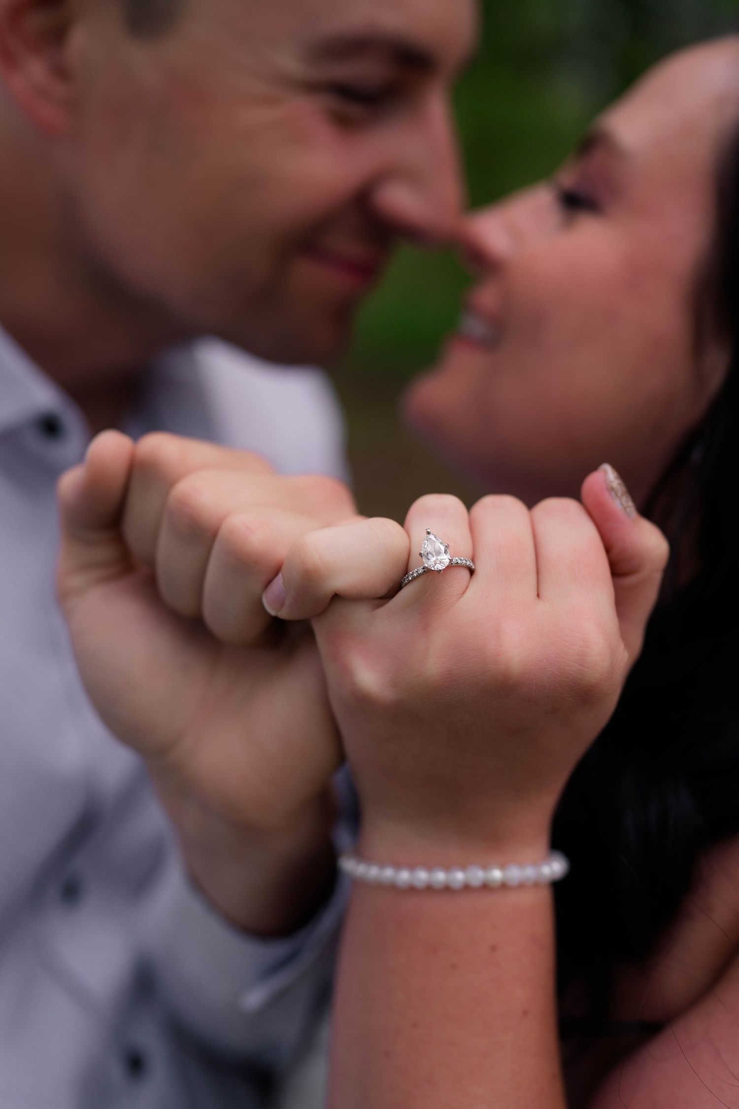 Close-up of engagement ring with pear-shaped diamond as smiling couple touches noses romantically.