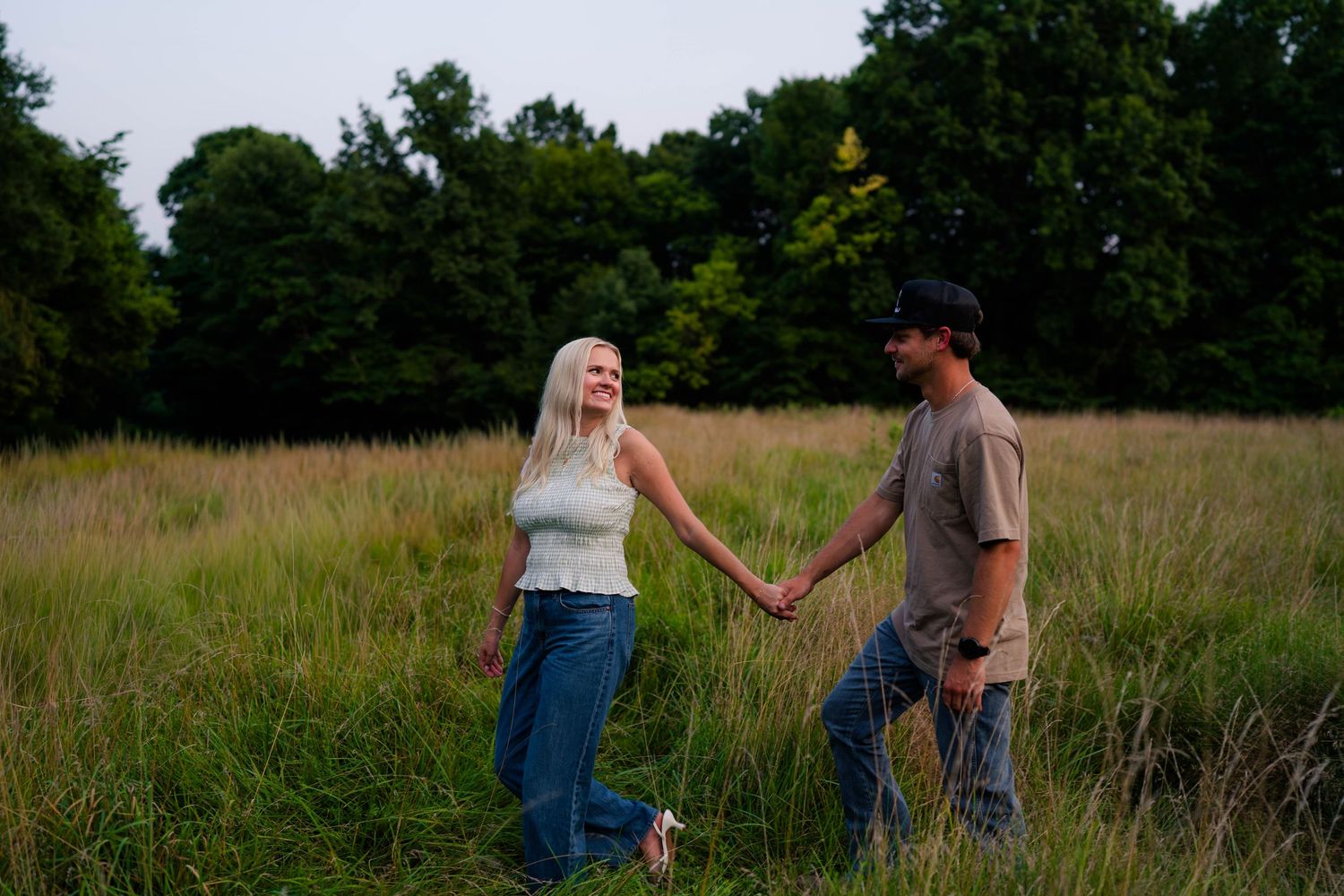 A couple holds hands walking through a tall grass field surrounded by lush green trees at dusk.