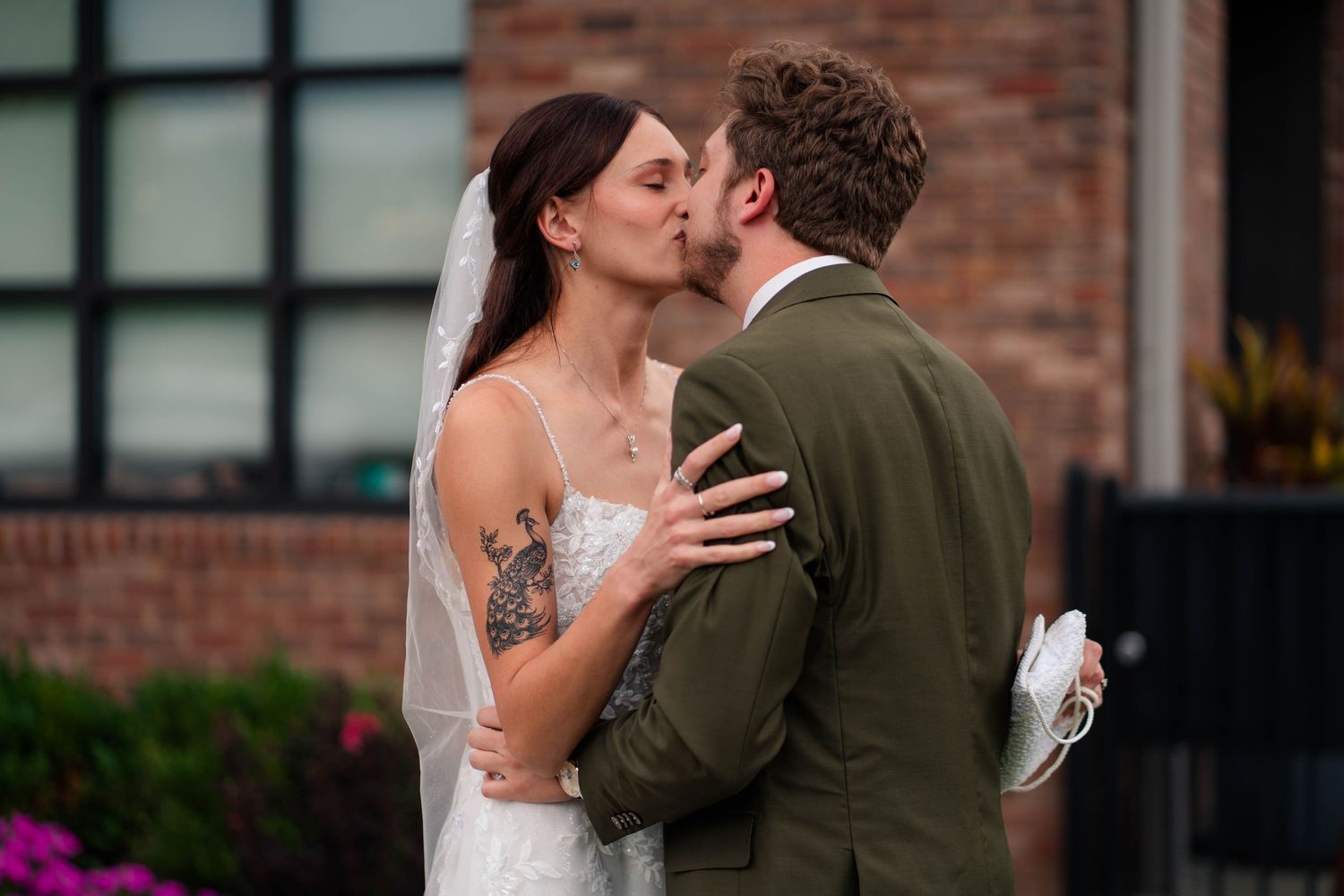 A bride and groom share a romantic kiss outdoors near a brick building on their wedding day.