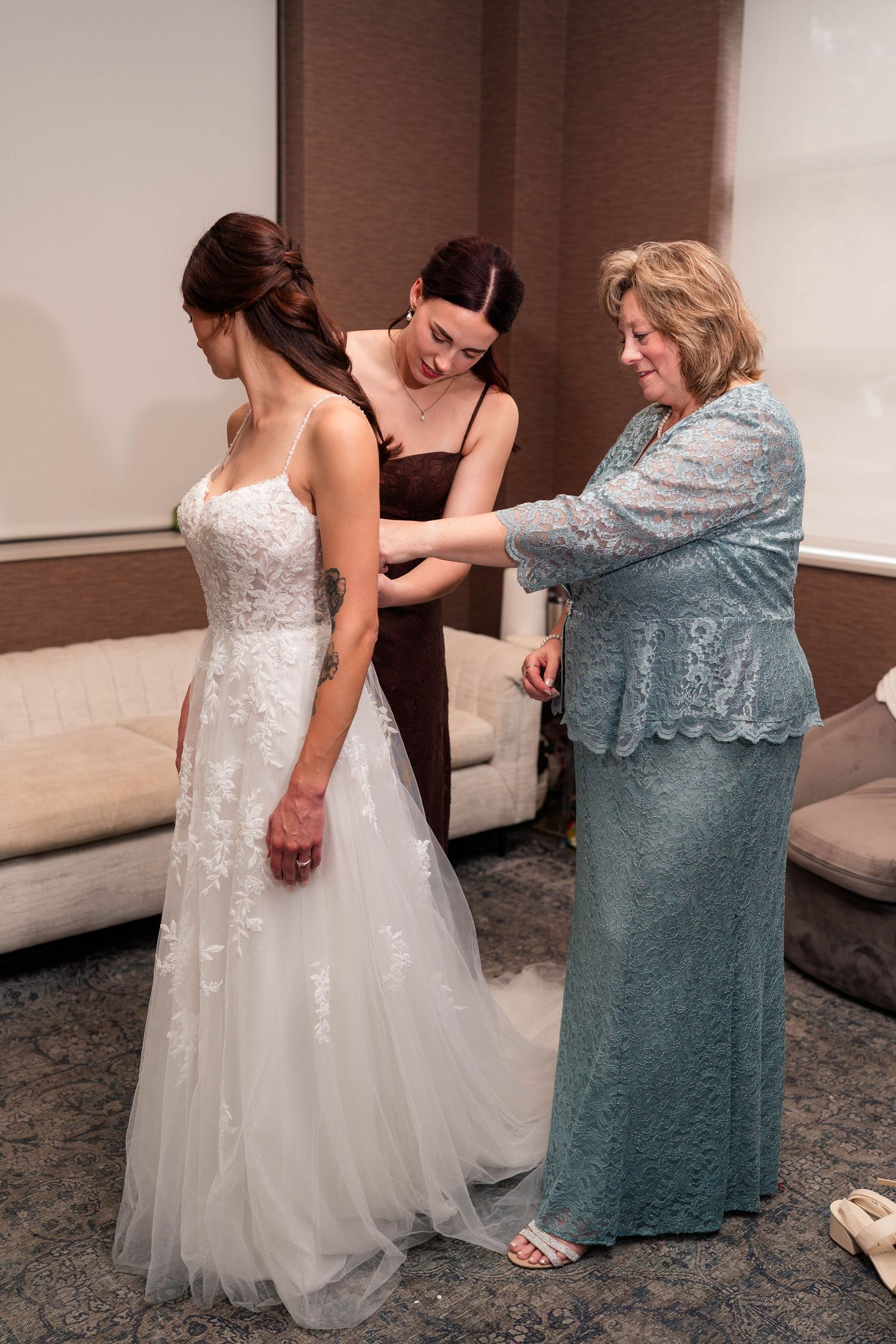 Mother and bridesmaid helping bride into her white lace wedding gown in a hotel bridal suite.