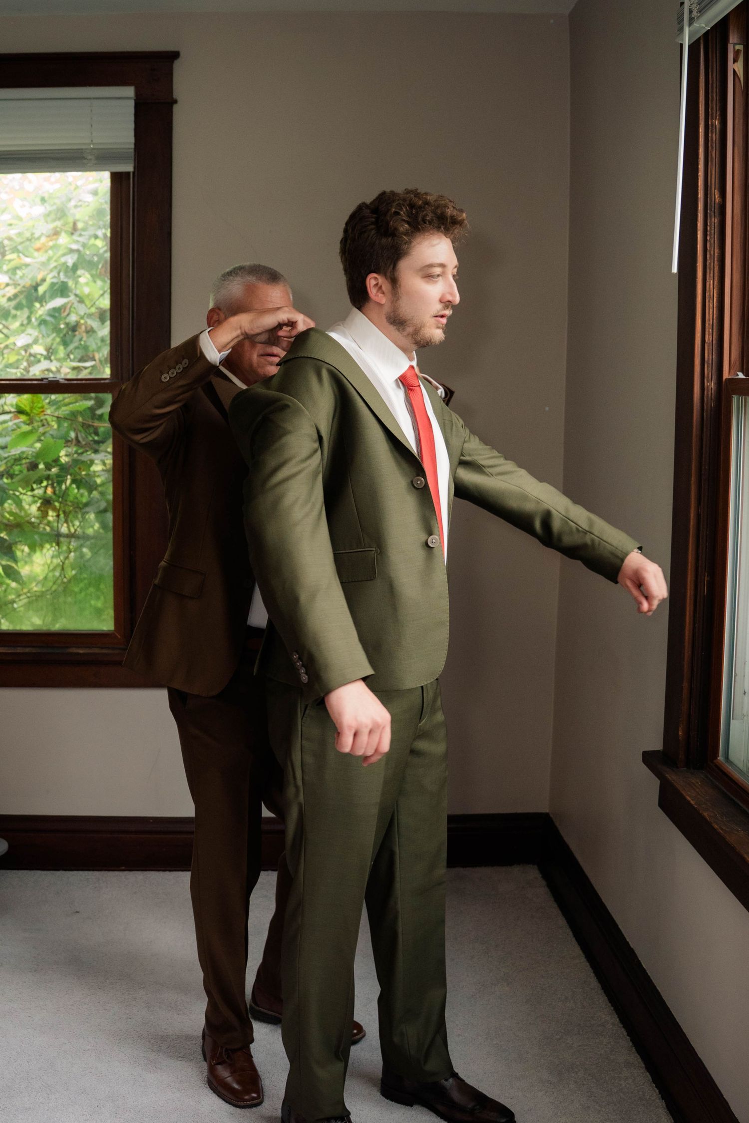 Two men in olive green suits with red ties stand near windows, one helping the other adjust his jacket.