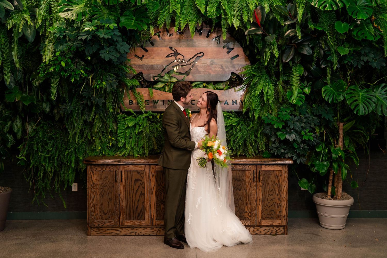 Bride and groom share a kiss in front of lush tropical green wall with wooden bar backdrop.