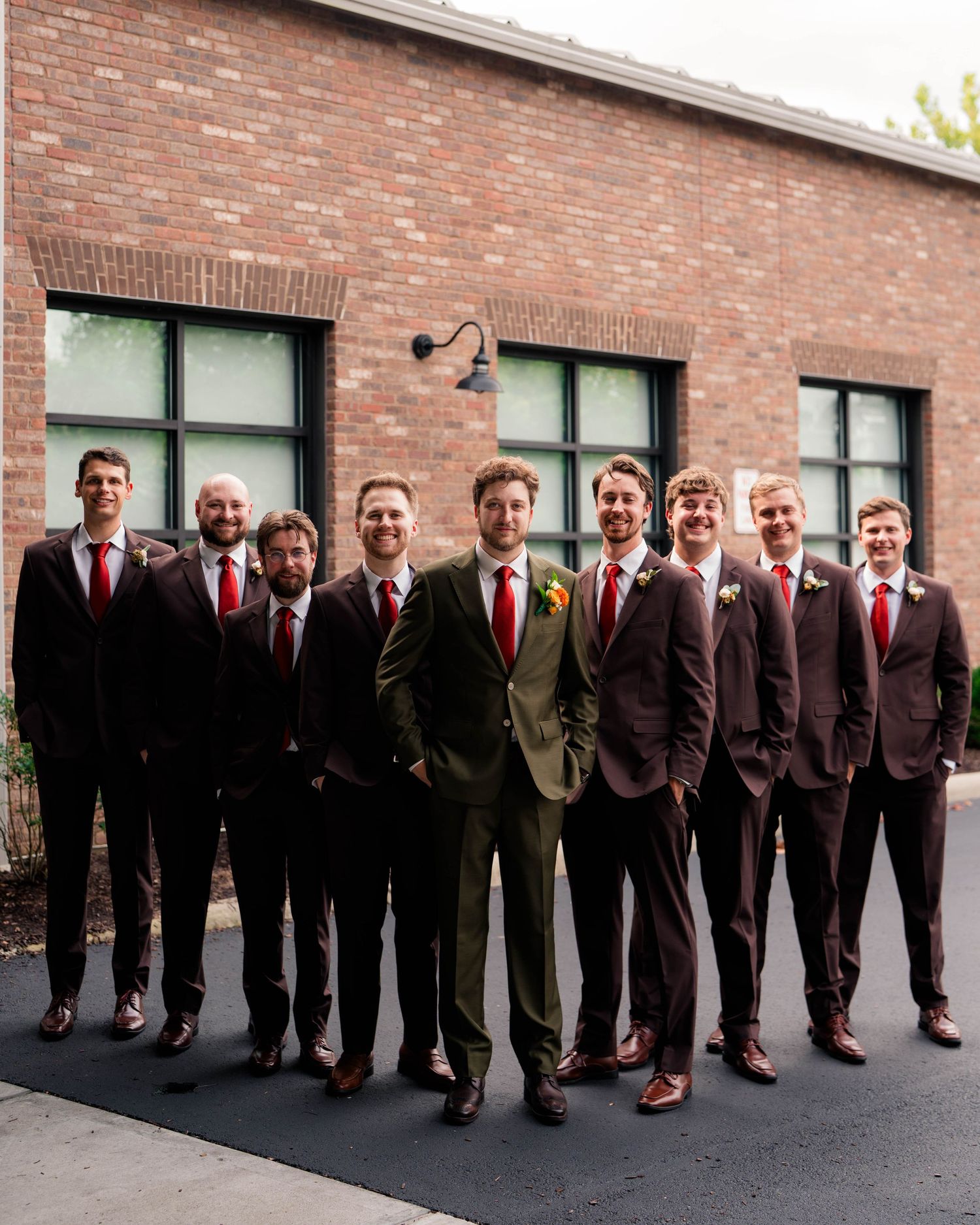 Groom in olive suit stands with groomsmen in dark brown suits and red ties outside brick building.
