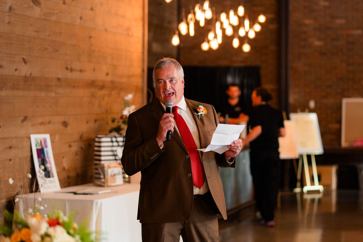 Wedding guest in brown suit and red tie giving heartfelt speech with microphone at reception.