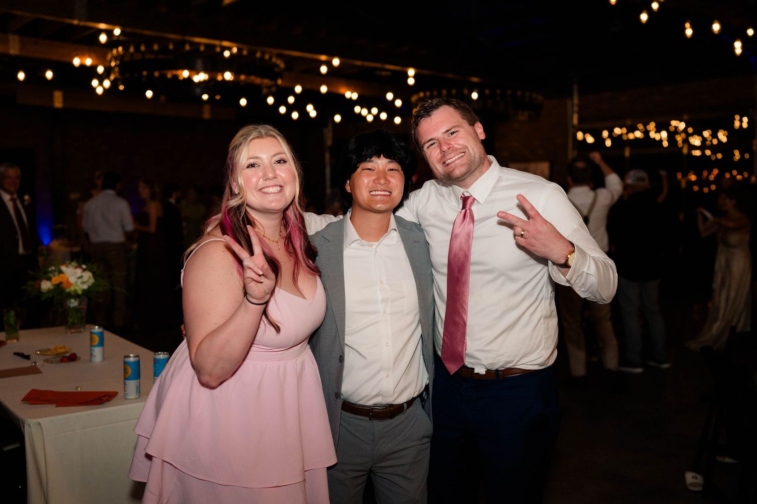 Three wedding guests pose and smile together under string lights at reception.