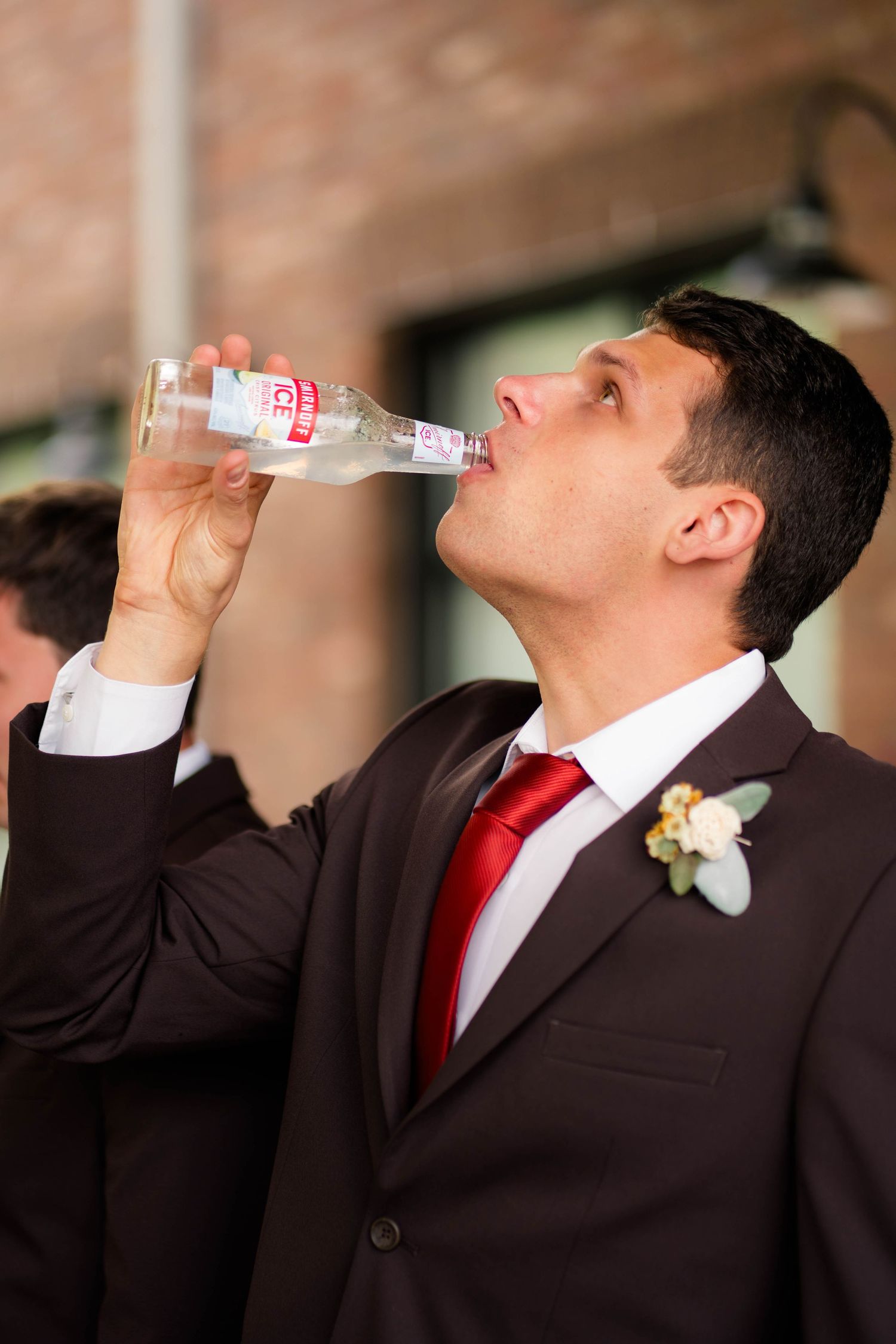 Groomsman in dark suit and red tie drinking Smirnoff Ice bottle at wedding.