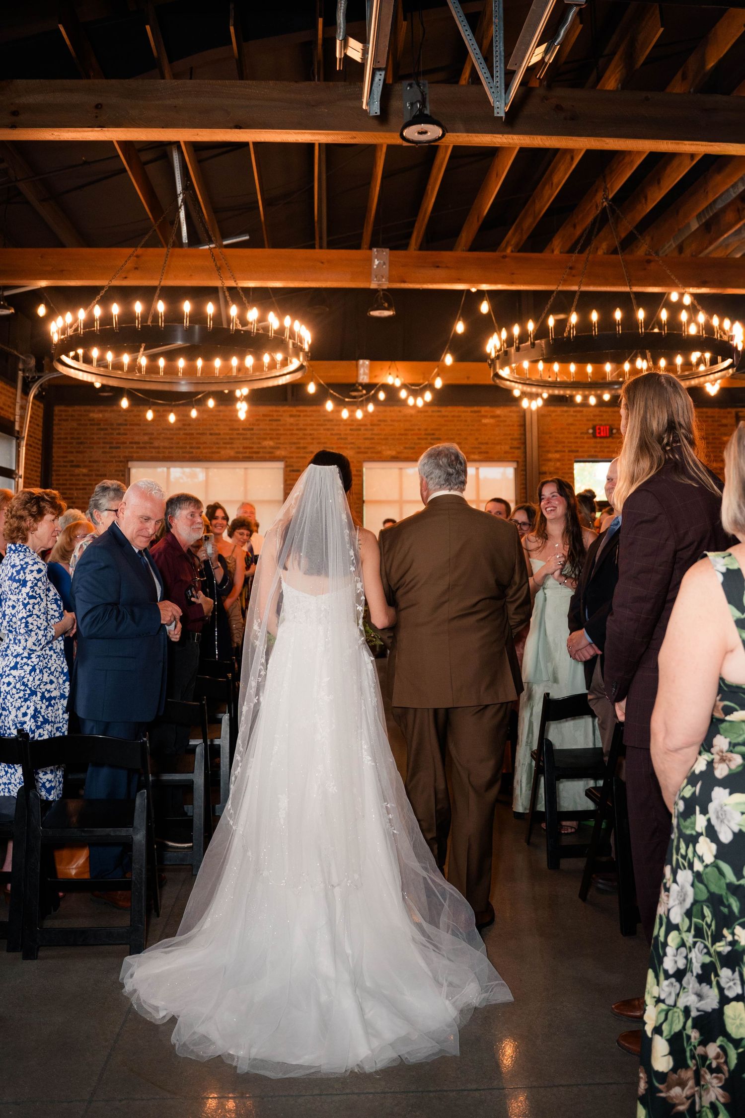 Bride in white gown and veil walks down the aisle with escort toward the altar in a warmly lit venue.