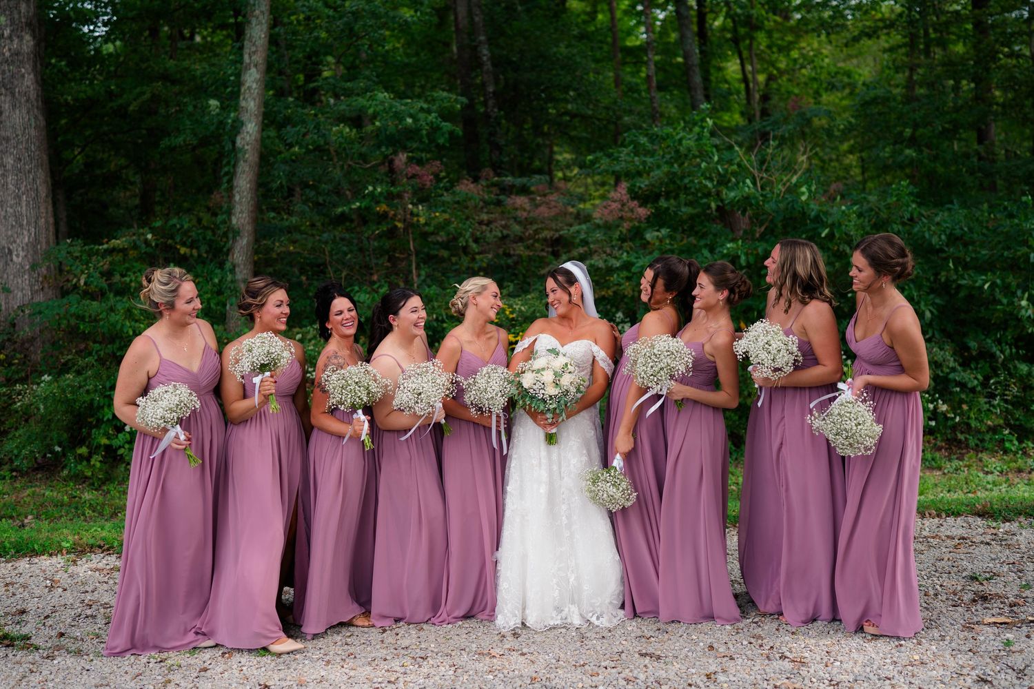 Bride stands with bridesmaids in mauve gowns holding baby's breath bouquets in a wooded setting.